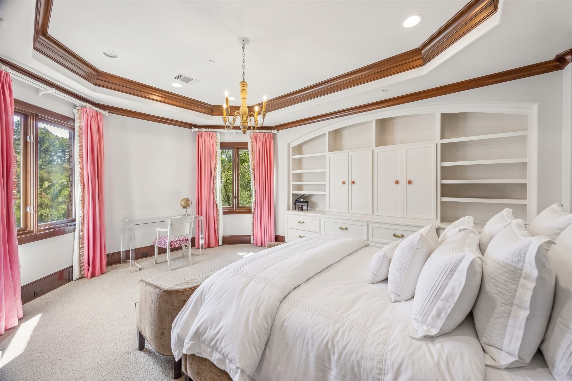 A bright upstairs bedroom with custom built-in shelving and cabinetry, detailed crown molding, and windows offering treetop views.