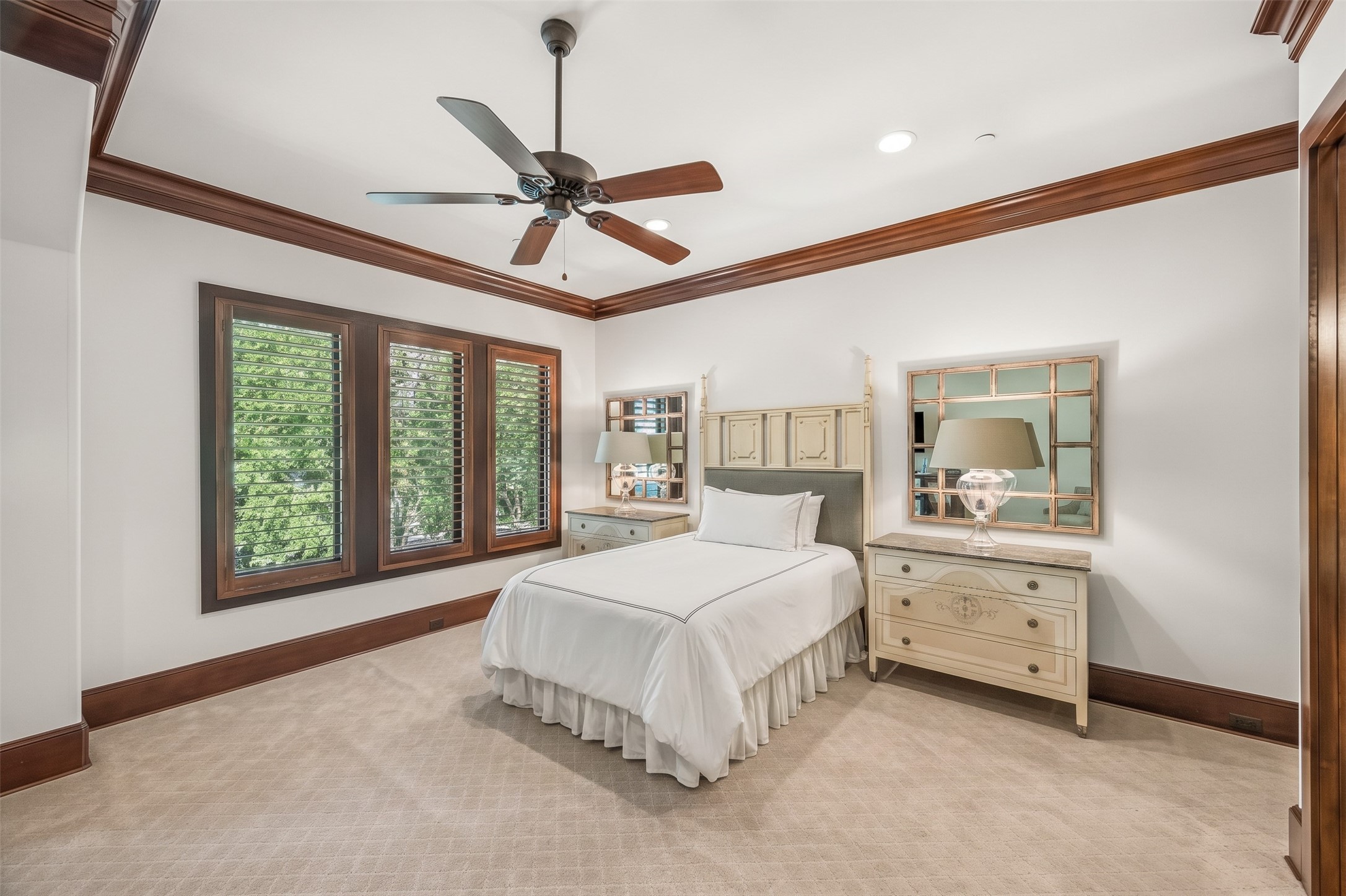 This upstairs bedroom offers a spacious layout with neutral carpeting, rich wood trim, and large windows with plantation shutters overlooking the surrounding trees.