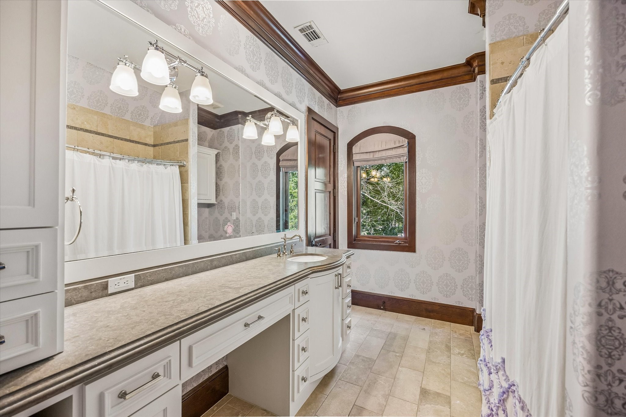 The adjoining bathroom offers a spacious vanity with generous counter space, custom cabinetry, a tub and shower combination, and elegant patterned wallcovering.