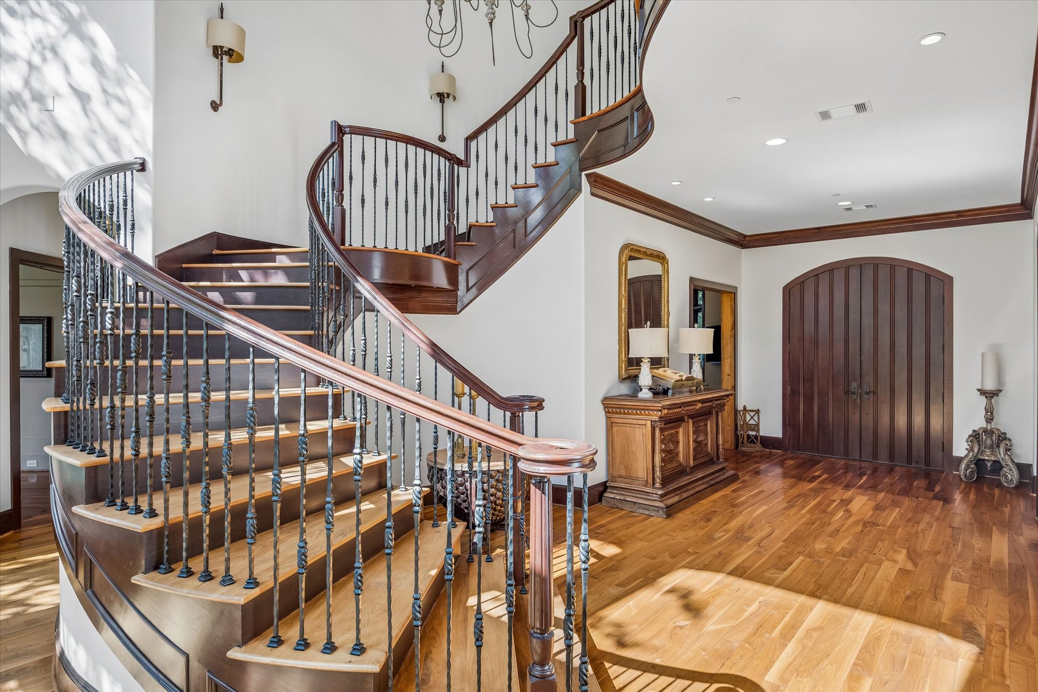 Grand two-story entry featuring a sweeping curved staircase with wood treads, wrought iron balusters, rich wood detailing, hardwood floors, and arched double entry doors.