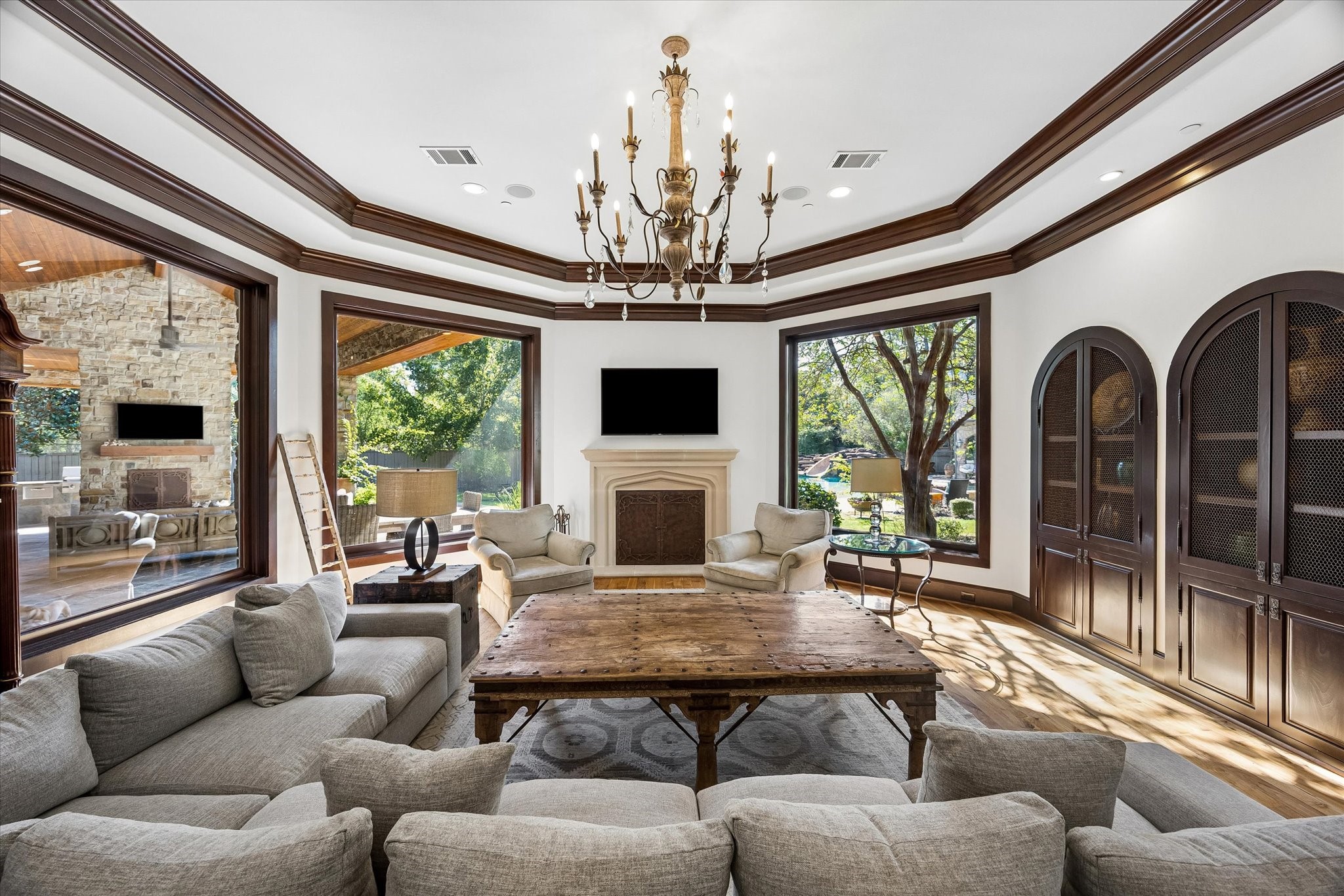 Another view of the family room highlights the custom arched cabinetry, detailed ceiling moldings, and expansive windows connecting the interior living space to the covered patio and pool beyond.