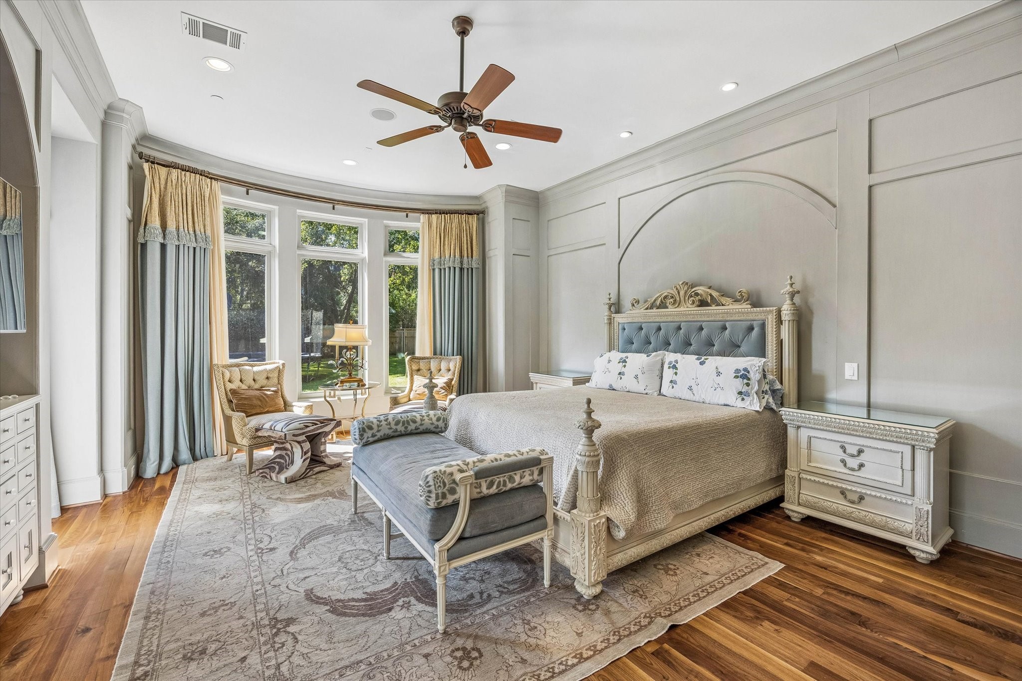 The first-floor primary suite offers a peaceful retreat with beautiful wall paneling, hardwood floors, a coffee bar, and a curved wall of windows overlooking the backyard.
