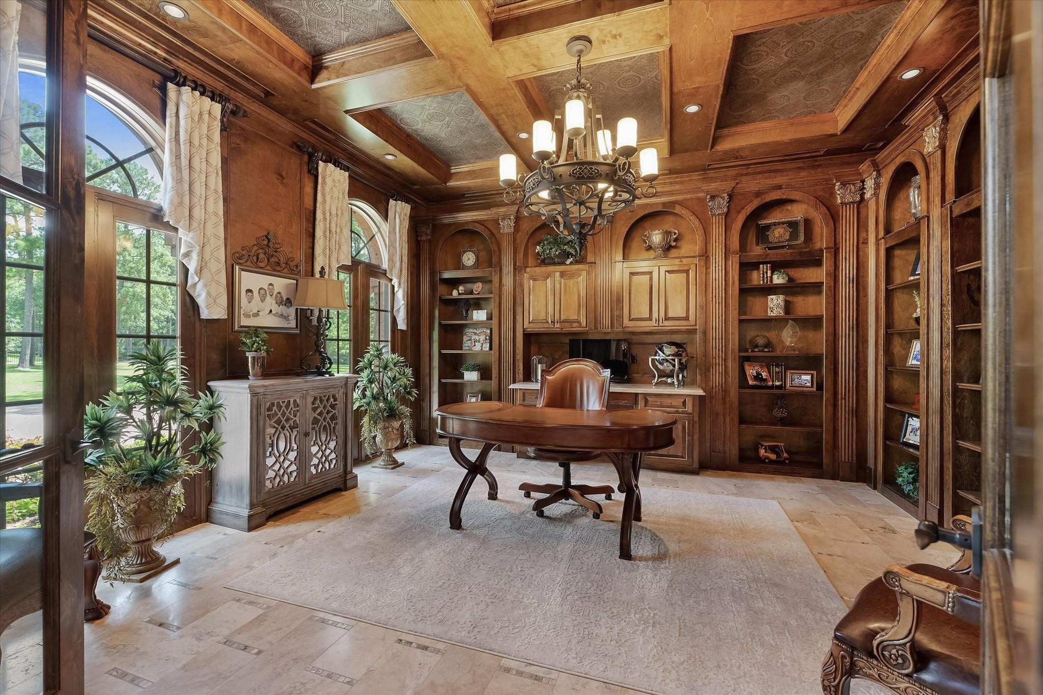 This study/library is fully paneled with mahogany wood and features extensive millwork on all walls.
It includes a built-in desk with custom cabinetry that offers lower closed storage and open shelving.
The room boasts a custom coffered tray ceiling with cove lighting, recessed lighting, a designer chandelier, and custom crown molding. Windows provide views of the front lawn with northern exposure, adorned with custom window coverings.
Access to the foyer is through French doors with a valence.