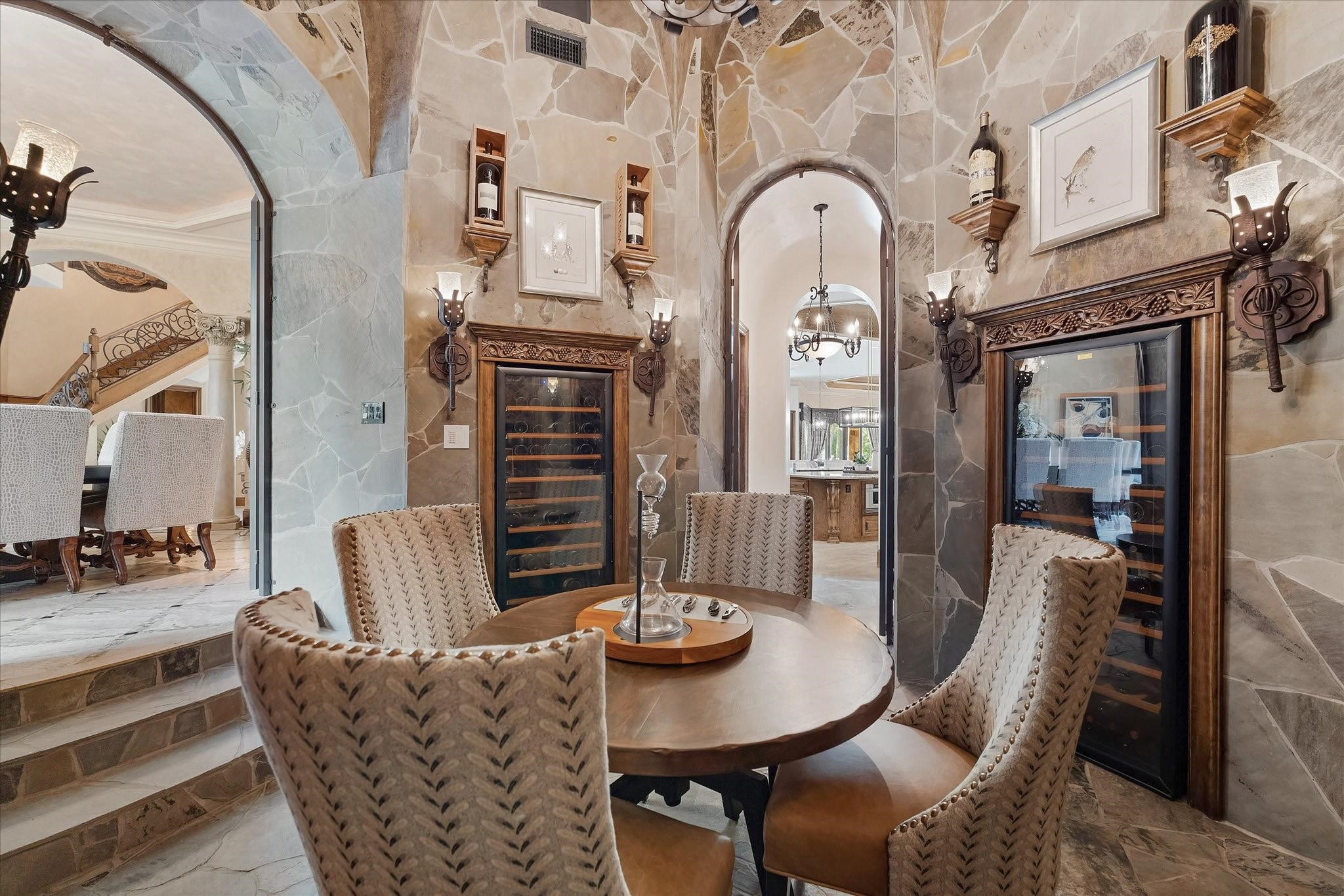 The Sunken wine cellar features two climate-controlled wine fridges surrounded by custom millwork and crown molding. The room is accessed through custom iron gate doors at both entryways and boasts stone floors and walls. Custom diamond pattern wine storage, marble countertops with lower storage and drawers, and vaulted cathedral ceiling. Illuminated by a custom chandelier, sconce torch lighting, and stained glass with internal lighting, offers storage for approximately 700 bottles.