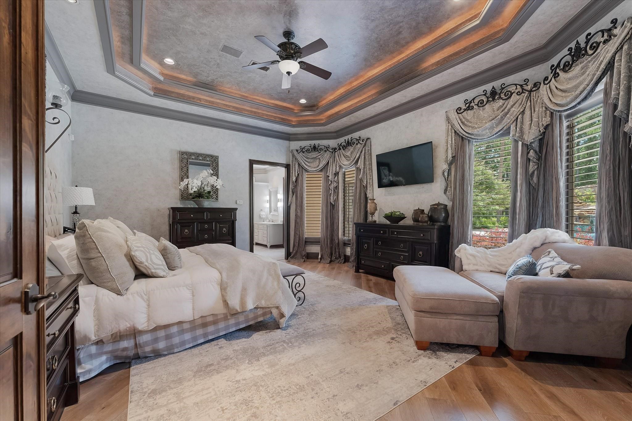 Located off the formal living room, the primary bedroom features a dual Adler wood door entry, recessed surround sound speakers, and a custom tray ceiling with cove lighting. Large windows offer views of the backyard with southern and eastern exposures. The room is adorned with custom designer window coverings, recessed LED lighting, custom wood shutters, hardwood floors, designer neutral wall paint, and custom crown molding, creating an elegant and inviting space.