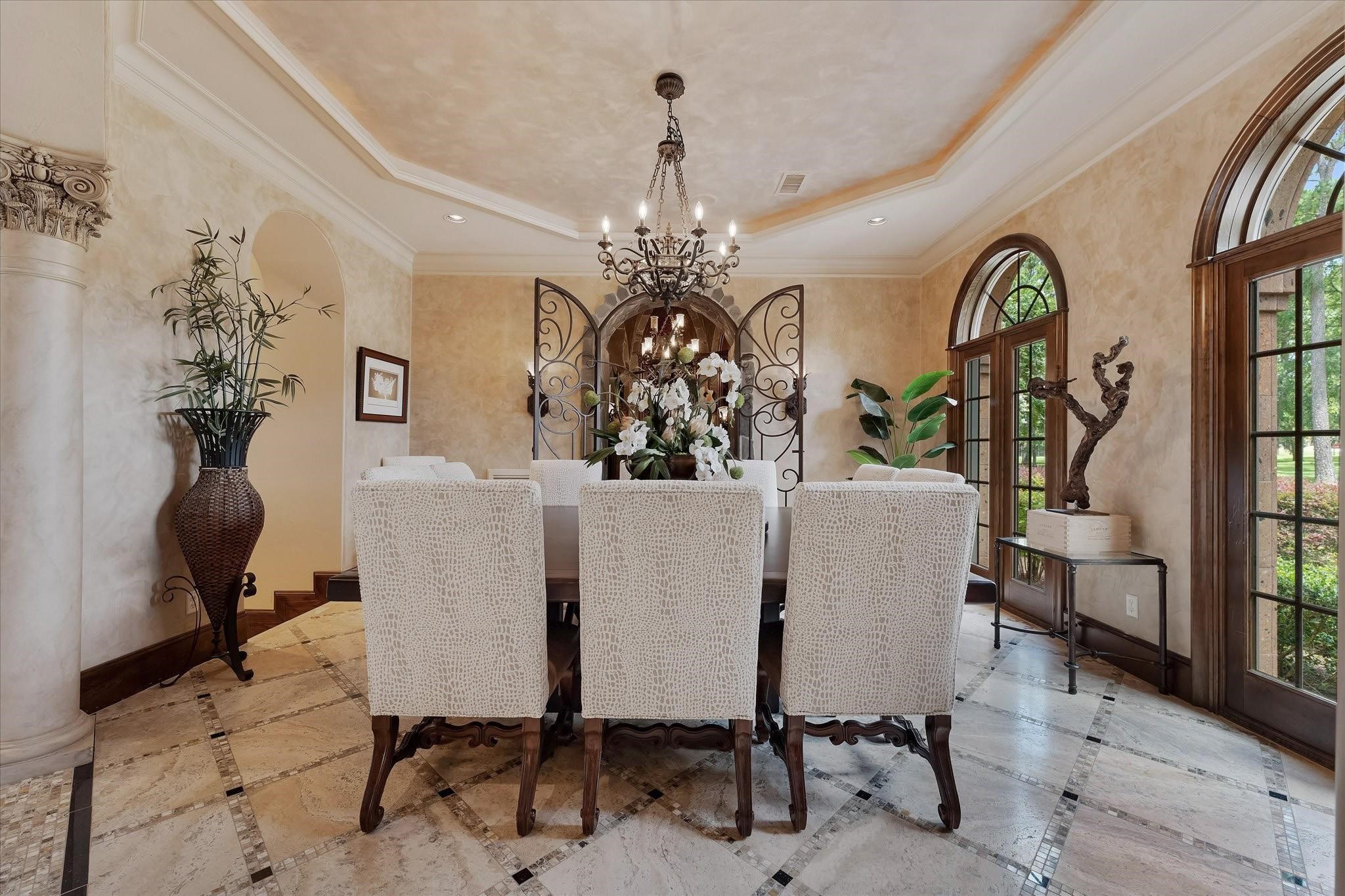 The formal dining room boosts arched columns and travertine flooring with a custom tile pattern.
Arched windows overlook the front lawn. Integrate crown molding, recessed speakers, recessed lighting, and a custom chandelier enhance the elegance. The custom tray ceiling with cove lighting and Venetian plastered walls add to the sophisticated ambiance. Custom arched iron gates lead to the wine room, and an arched opening leads to the butler's pantry hallway, providing two different ways to access the kitchen