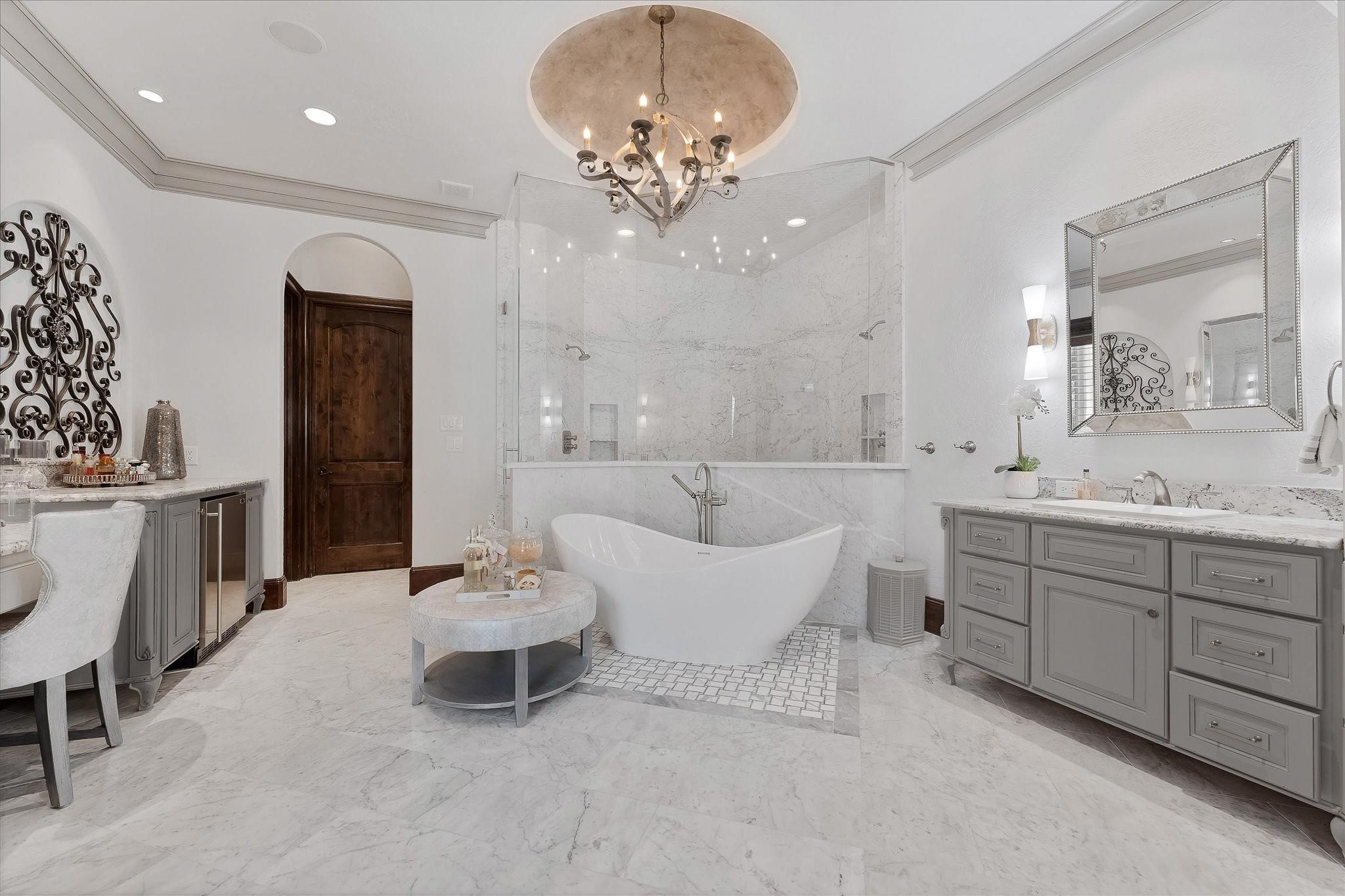 Fully renovated Primary ensuite boosting Carrara marble walk-in steam shower with a frameless glass entry door, dual shower heads, designer polished nickel plumbing fixtures, built-in bench seats, and Carrara marble mosaic tile on the floor and ceiling. A freestanding soaking tub surrounded by Intricate tile pattern and Carrara marble flooring. An exterior door with custom wood shutters leads to the backyard and pool area,