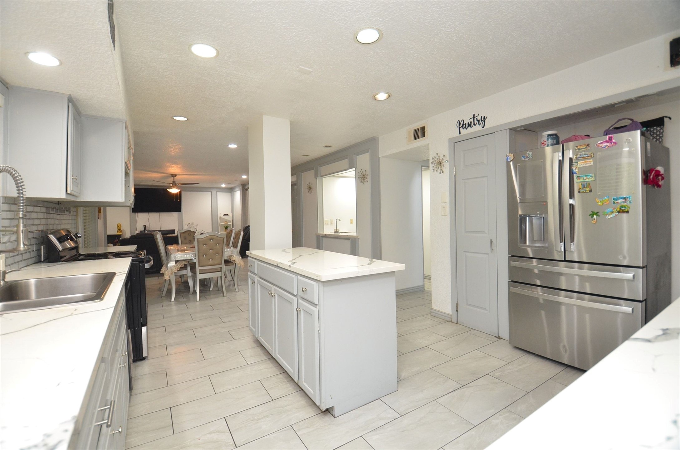 Open-concept kitchen with center island featuring stylish modern quartz tops, painted gray cabinetry, pantry closet, stainless-steel refrigerator, recessed lighting and light tile flooring, flowing seamlessly to the dining and living areas.