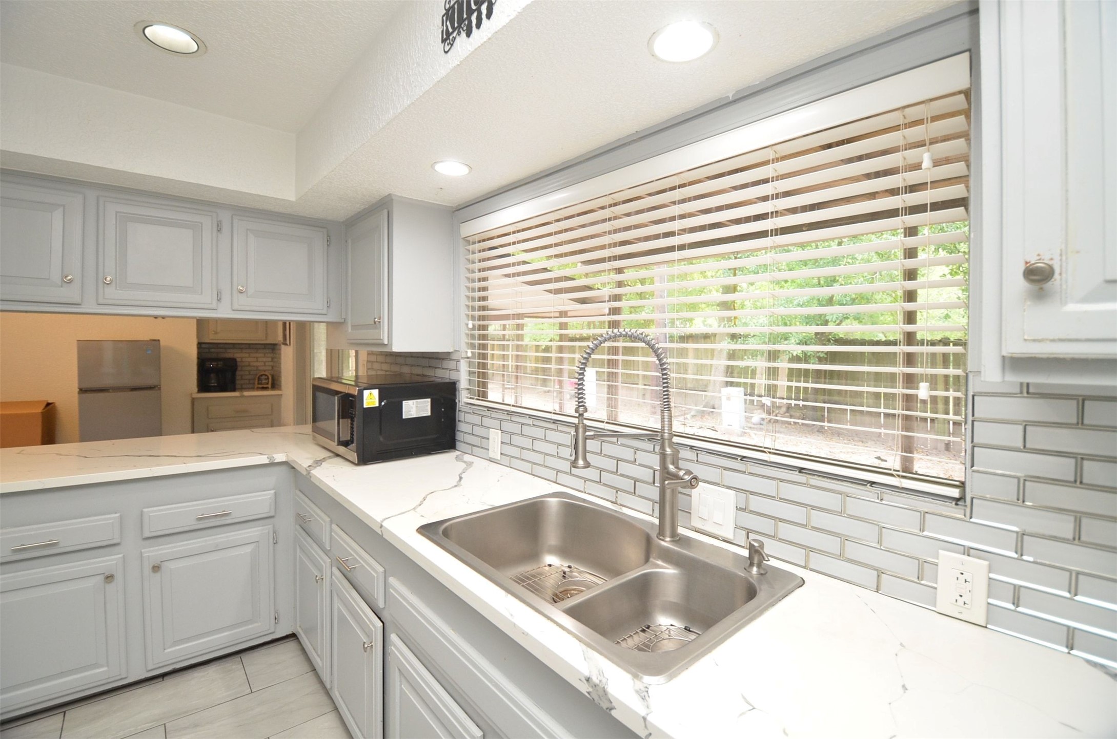 New open-concept kitchen with stylish modern quartz counters, painted gray cabinetry, subway-tile backsplash, stainless double sink with gooseneck faucet, recessed lighting, and large window overlooking the backyard.
