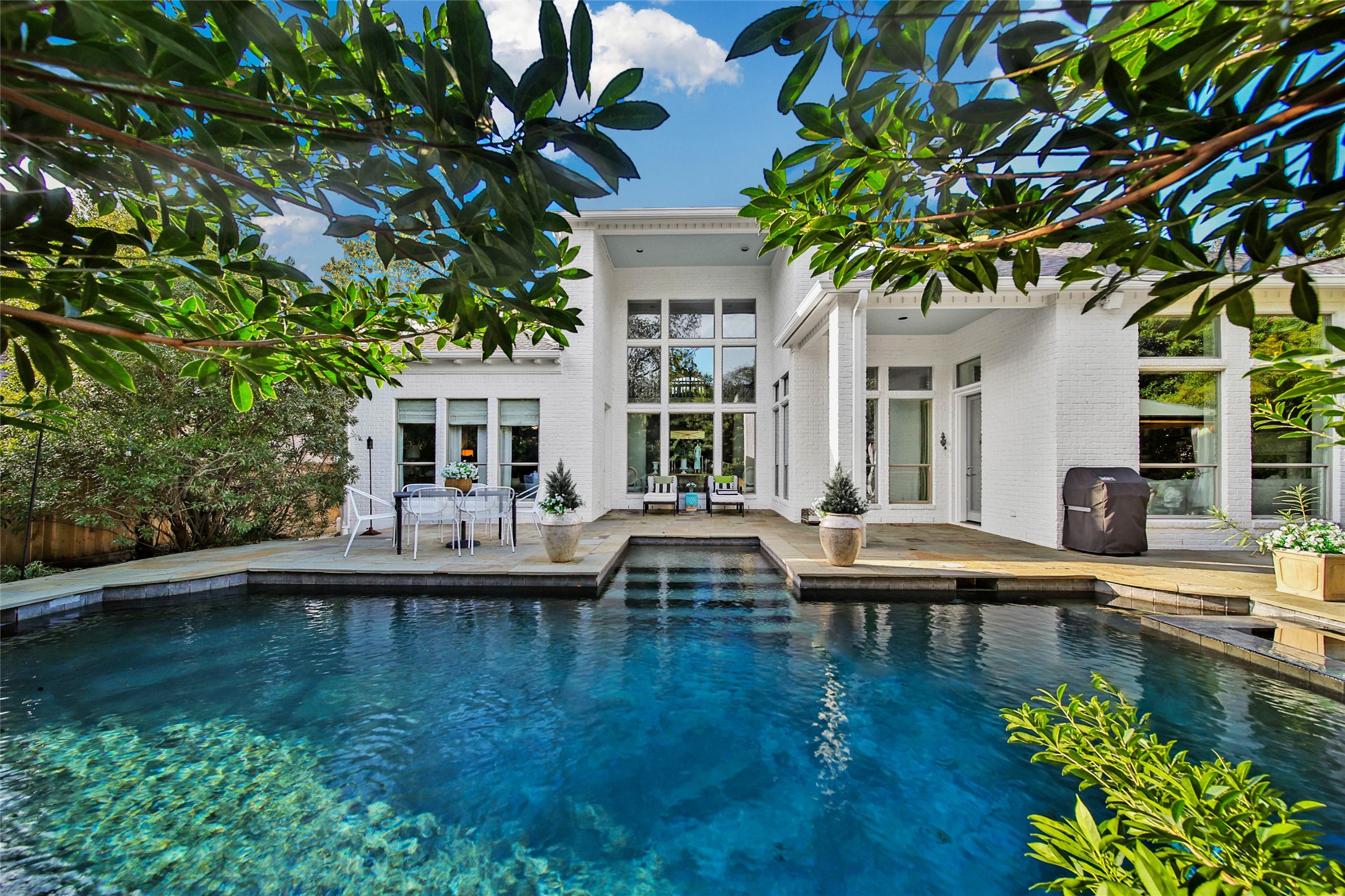 Classic home with a luxurious pool, patio, and abundant natural light from large windows, all surrounded by lush greenery.