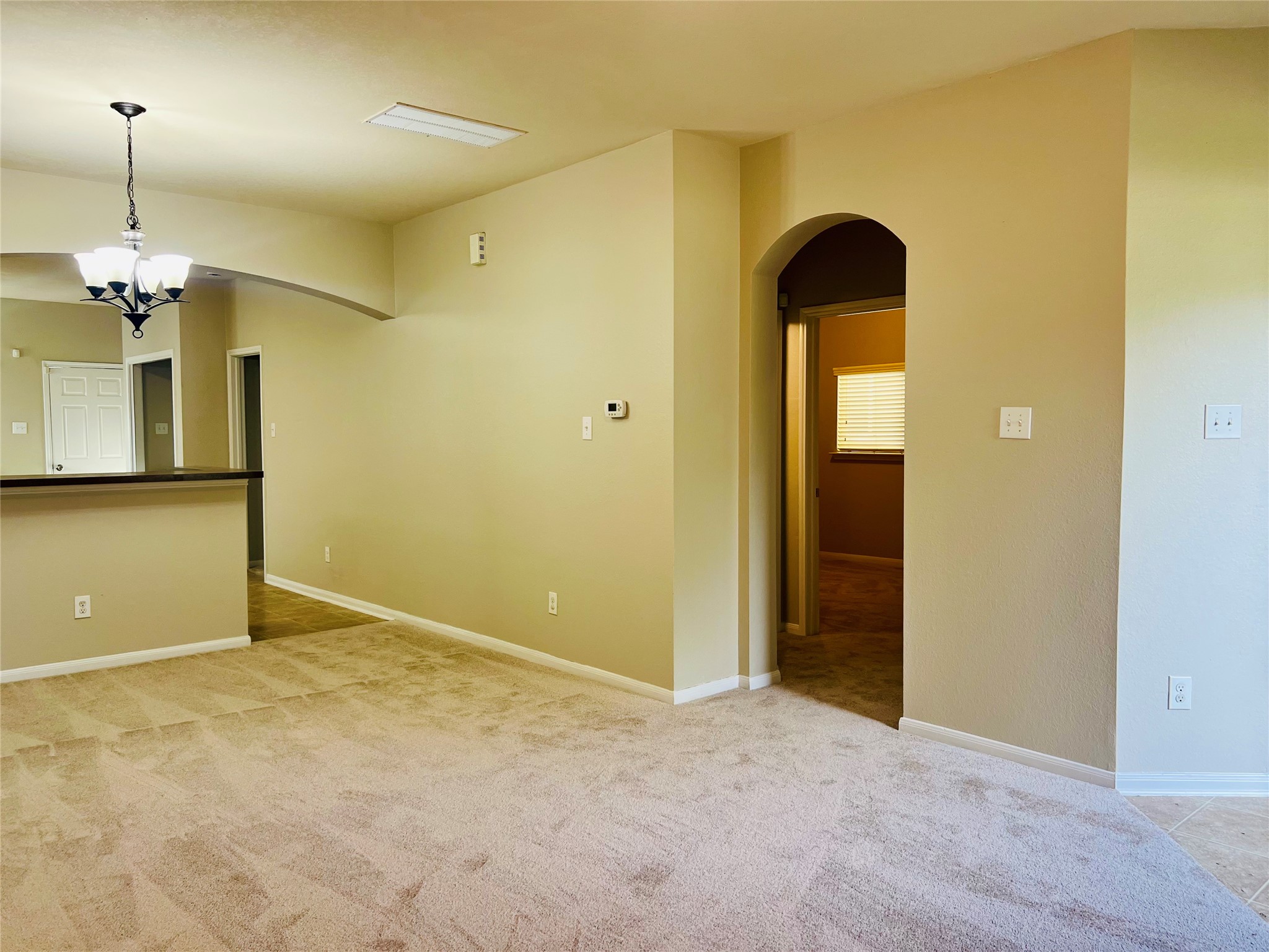 Spacious living area with plush carpet flooring, warm neutral walls, and a charming arched doorway leading to additional rooms. The open layout includes a modern light fixture and a partial view of the kitchen with a high countertop.