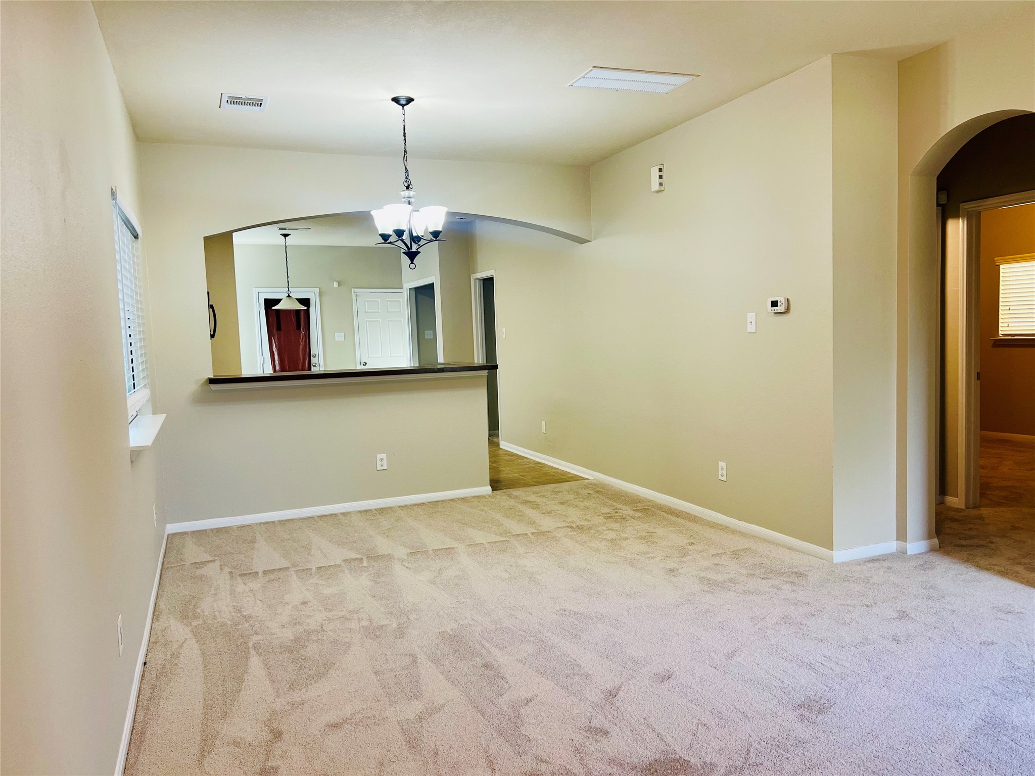 Spacious living area with plush carpeting, neutral walls, and an elegant arched opening leading to the kitchen, featuring modern light fixtures and a convenient breakfast bar.