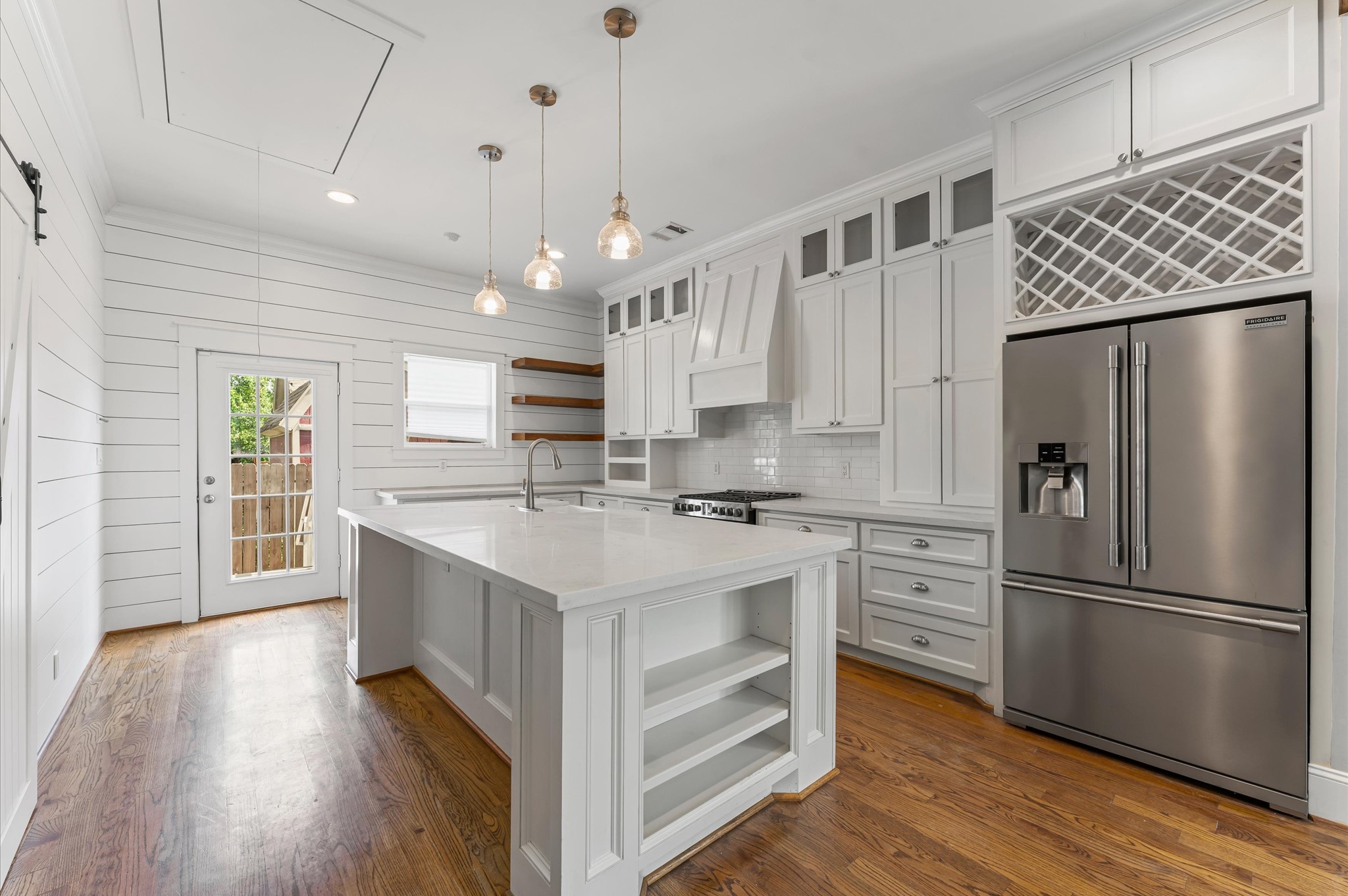 This modern kitchen features a spacious layout with white cabinetry, a large center island, stainless steel appliances, and elegant pendant lighting. The room is complemented by hardwood floors and a door leading to the outside, offering ample natural light.
