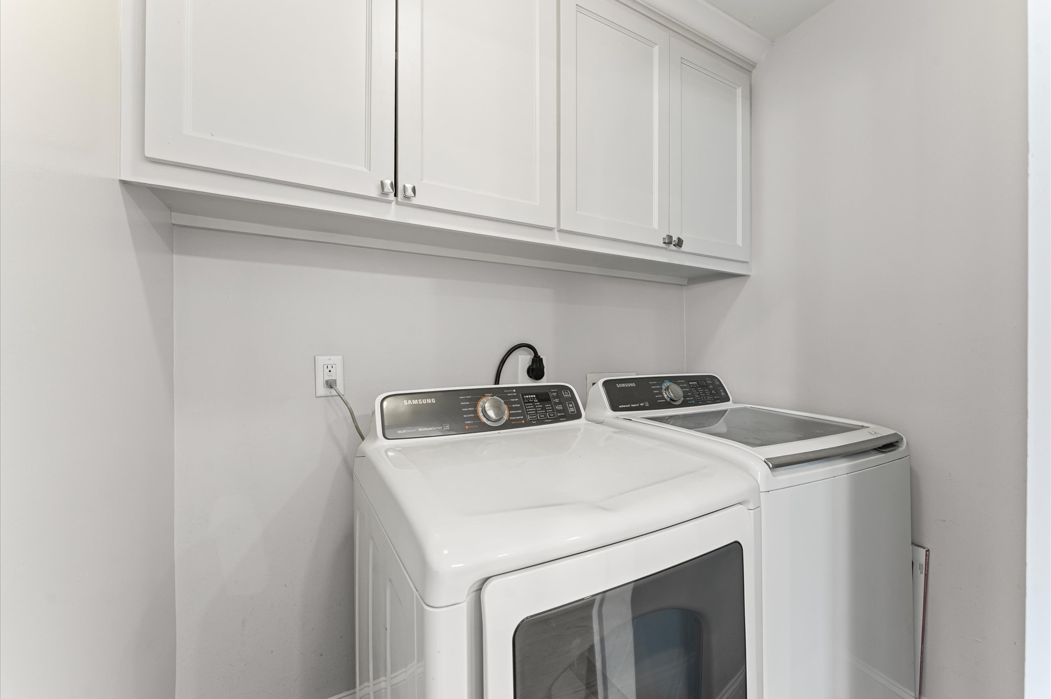 This photo showcases a compact laundry room with a modern washer and dryer set, featuring overhead white cabinets for storage. The space is clean and efficiently organized.