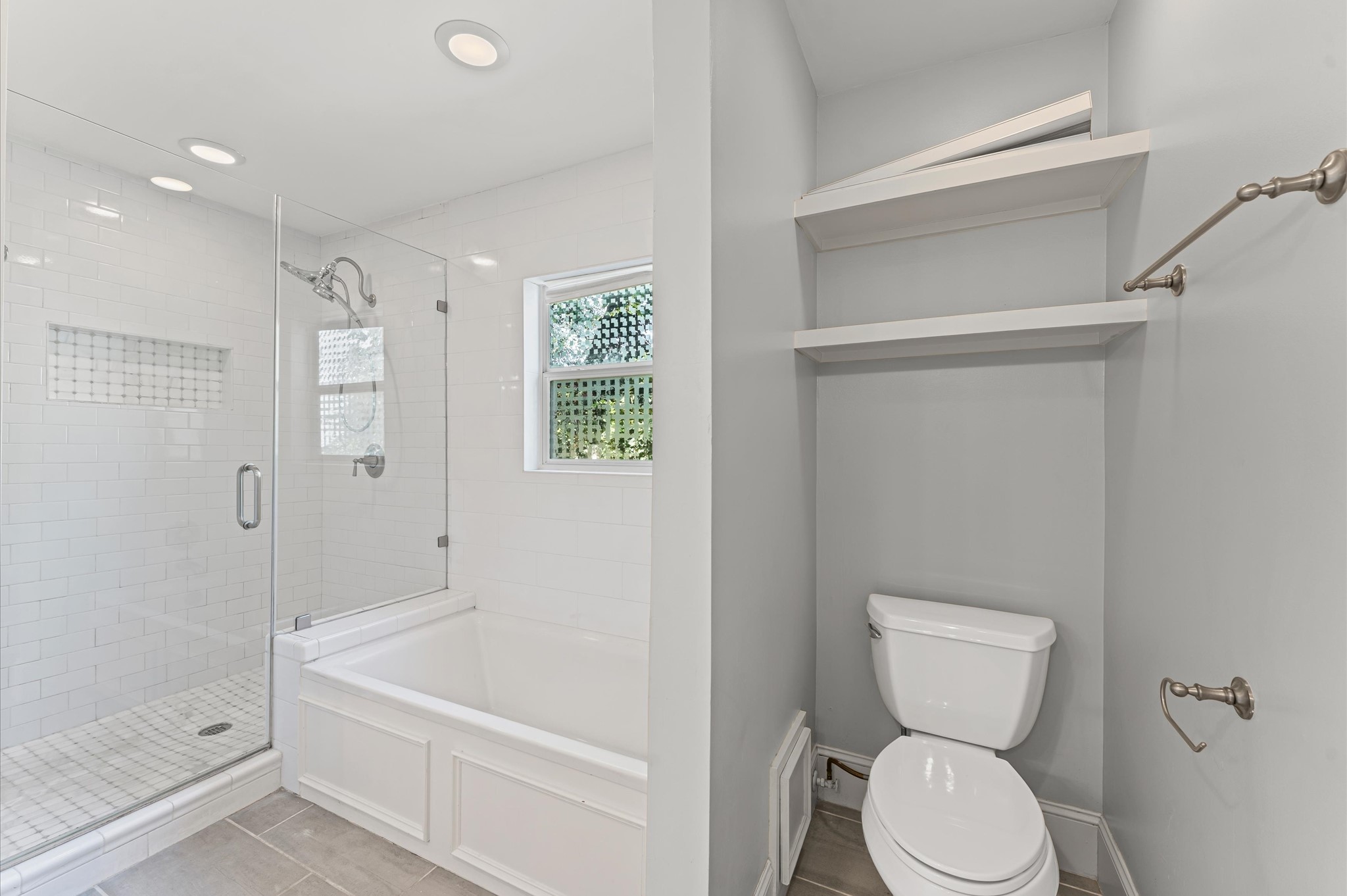 Modern bathroom featuring a glass-enclosed shower and bathtub combo, with subway tile walls. There's a small window for natural light, a toilet with shelving above, and sleek fixtures.