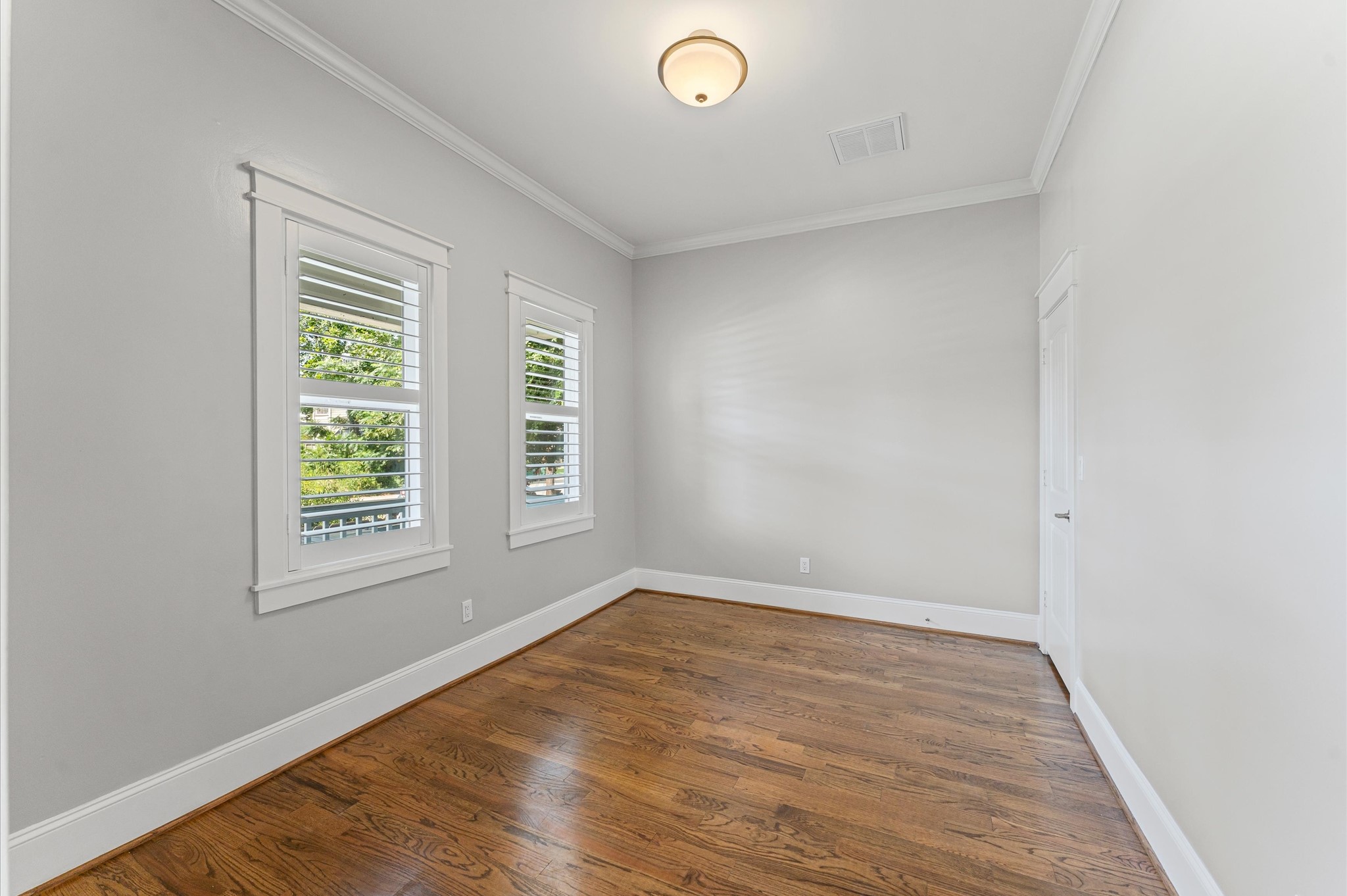 Bright room with hardwood floors, light gray walls, and crown molding. Features two windows with shutters, a ceiling light, and a white door. Ideal for a bedroom or office.