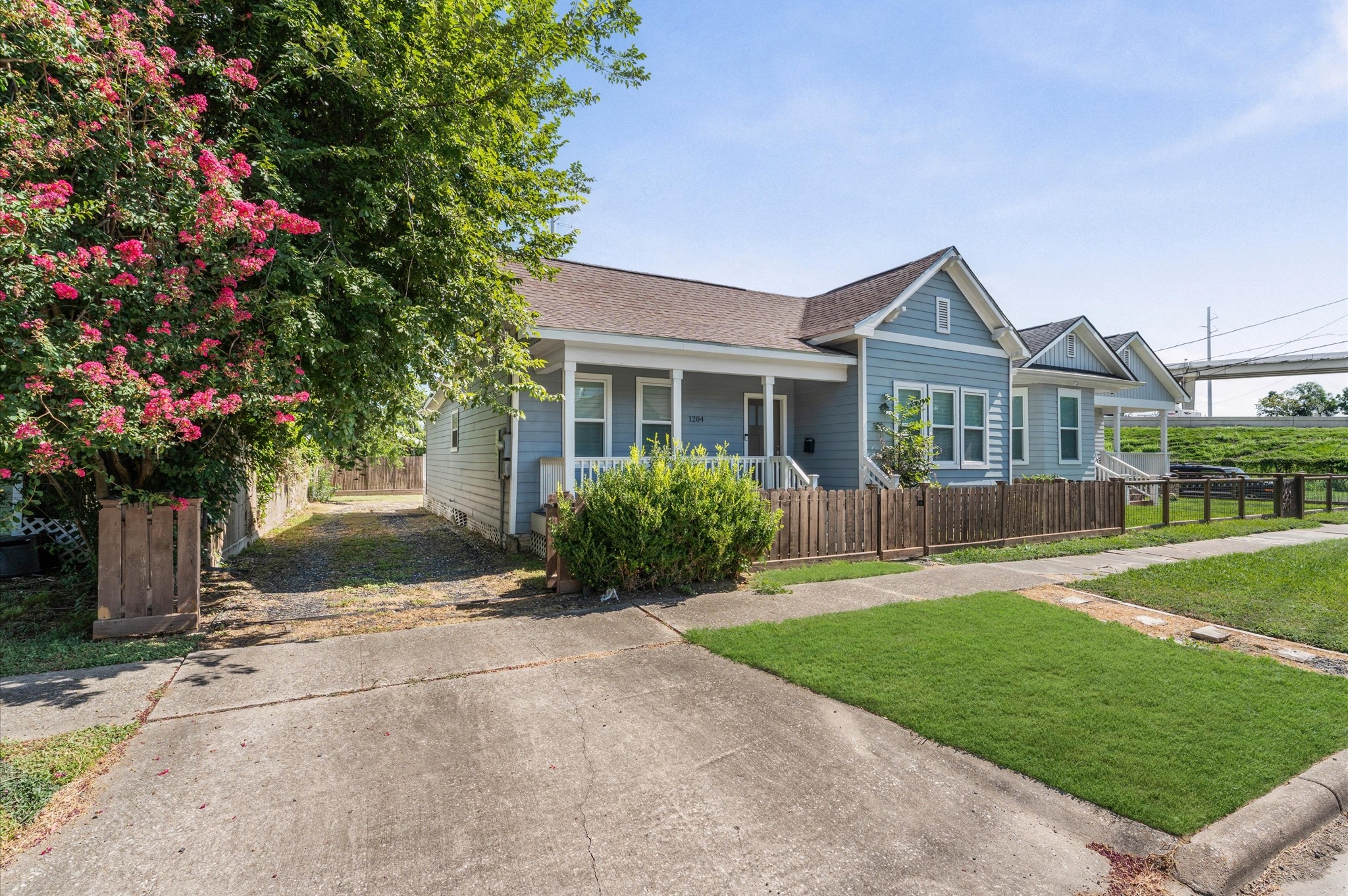 Charming blue bungalow with a welcoming front porch, surrounded by a wooden fence and lush greenery. The property features a well-maintained lawn, vibrant flowering trees, and a paved driveway, offering a cozy, inviting atmosphere.