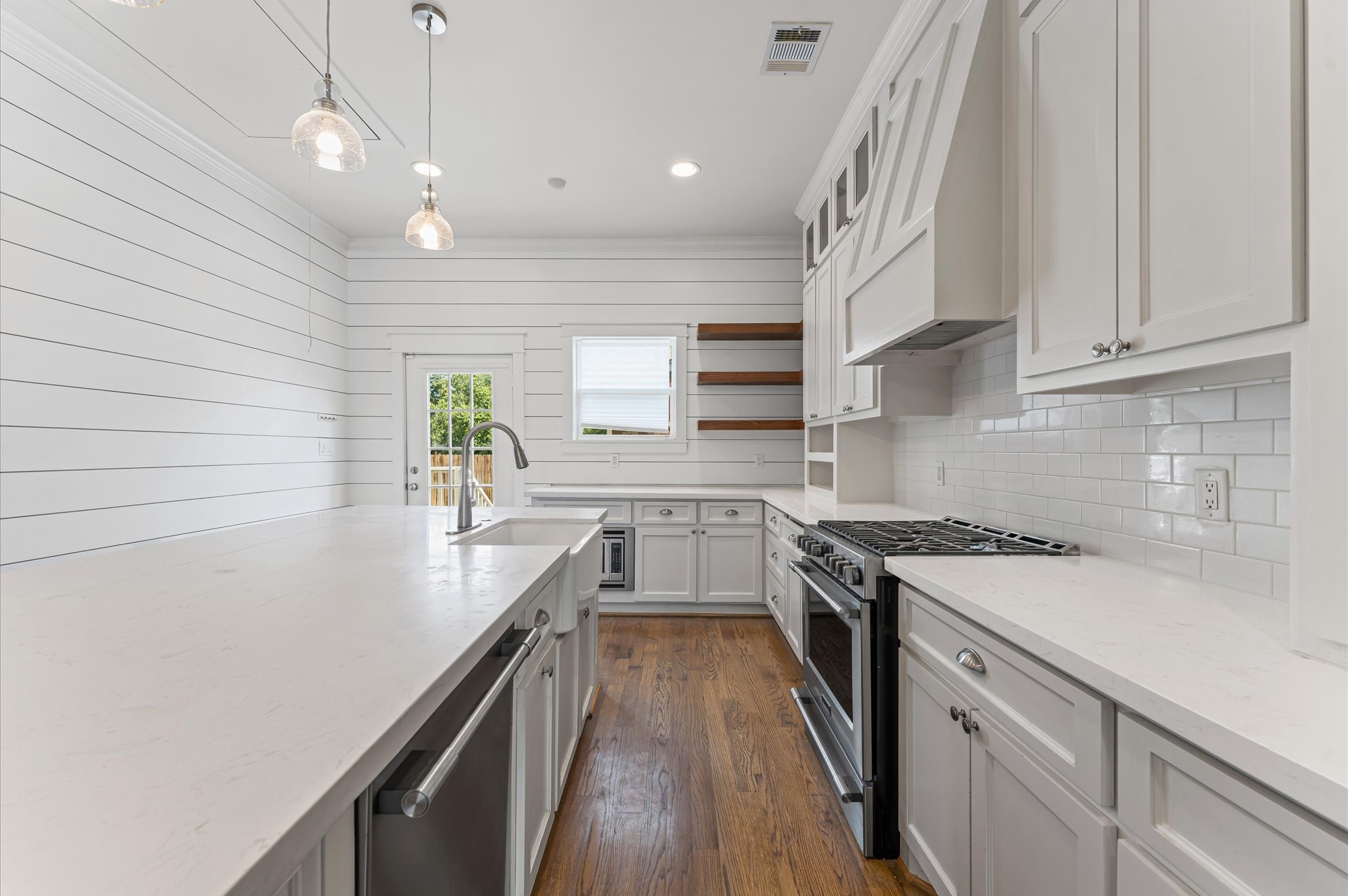 This kitchen features a bright, contemporary design with white cabinetry and shiplap walls. It includes modern stainless steel appliances, a farmhouse sink, and a spacious island with pendant lighting. The wooden flooring adds warmth, and there's ample natural light from a window and glass door.