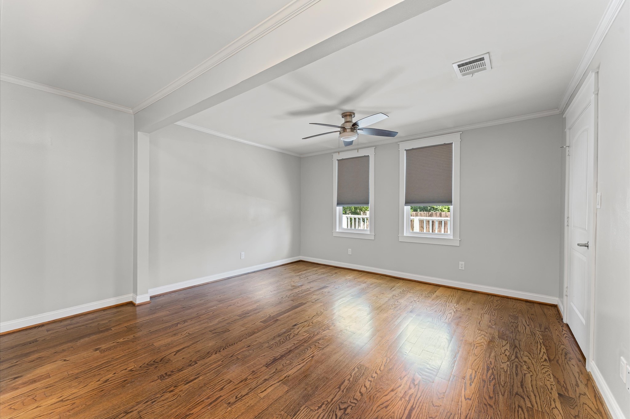 Spacious, bright room with hardwood floors, neutral walls, a ceiling fan, and two windows with blinds. Ideal for a bedroom or living space.