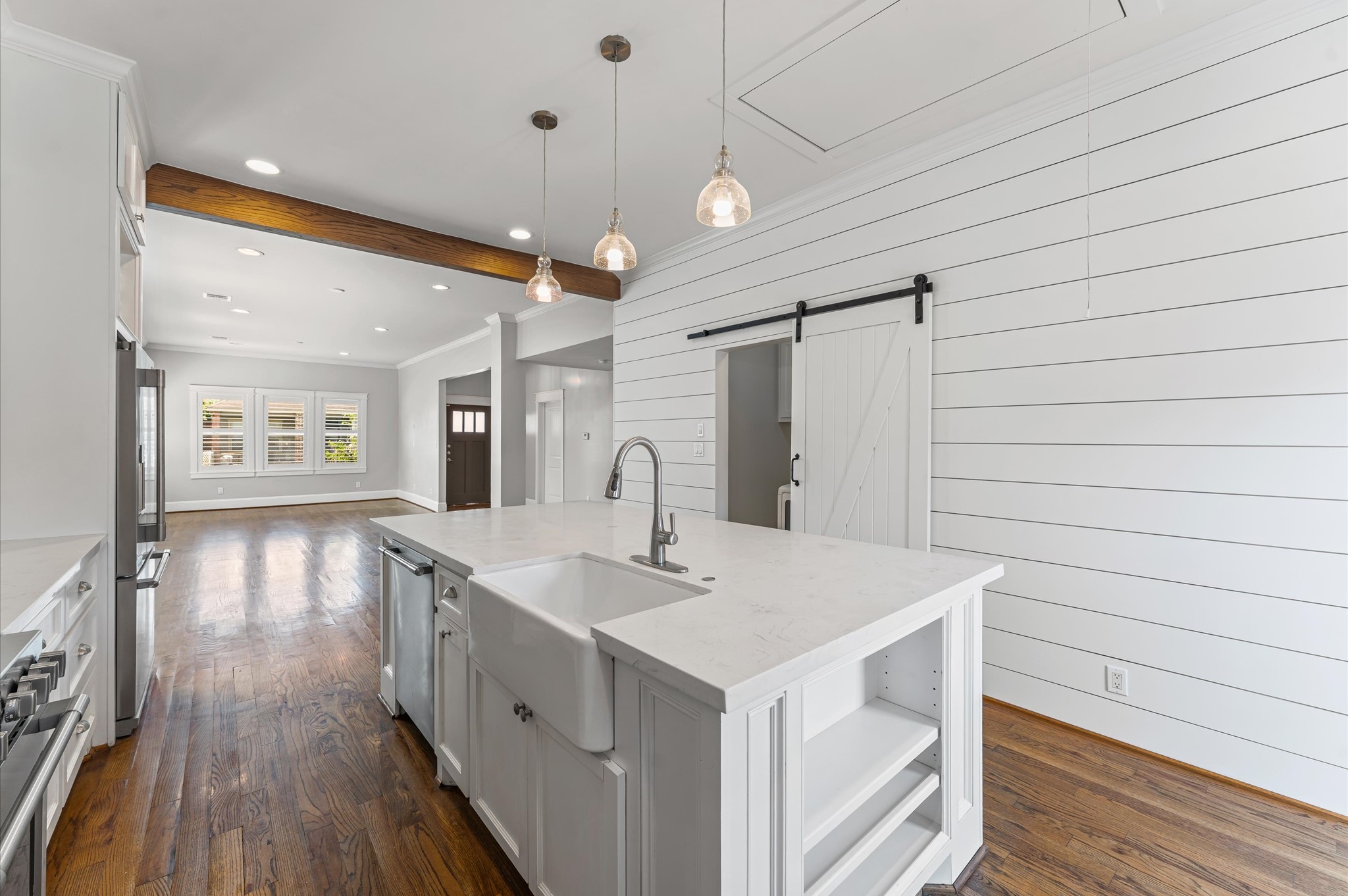 This bright kitchen features white shiplap walls, a large island with a farmhouse sink, and pendant lighting. It opens to a spacious living area with hardwood floors and a sliding barn door.