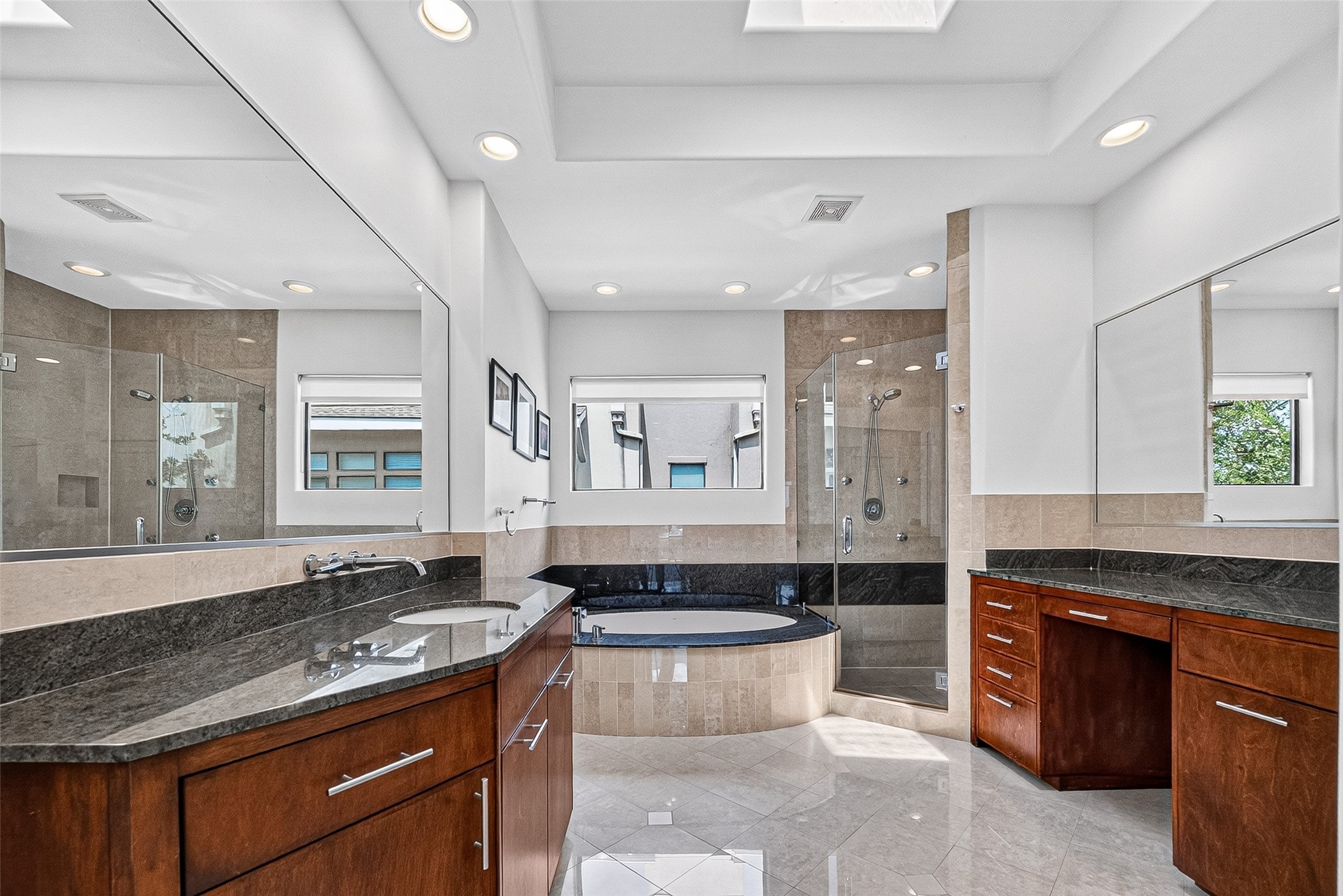 Luxurious primary bathroom featuring a spacious soaking tub, elegant granite countertops, wood cabinetry, and a glass-enclosed shower, all illuminated by natural light.