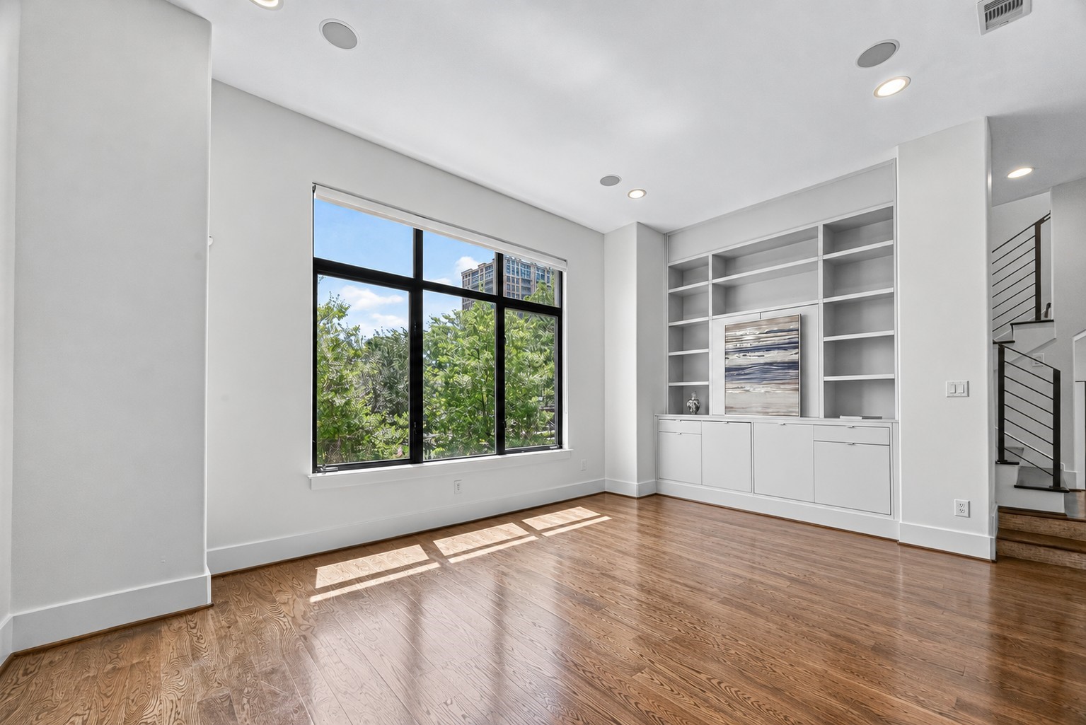 Sunlit living room with large windows showcasing lush greenery, elegant built-in shelving, and rich hardwood floors.