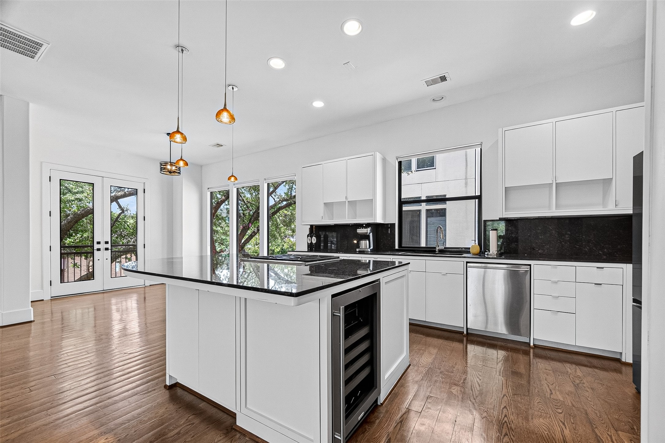 Elegant kitchen featuring sleek white cabinetry, a spacious island with a cooktop and wine cooler, and stunning hardwood floors. Abundant natural light streams in through large windows and glass doors, offering a view of lush greenery.