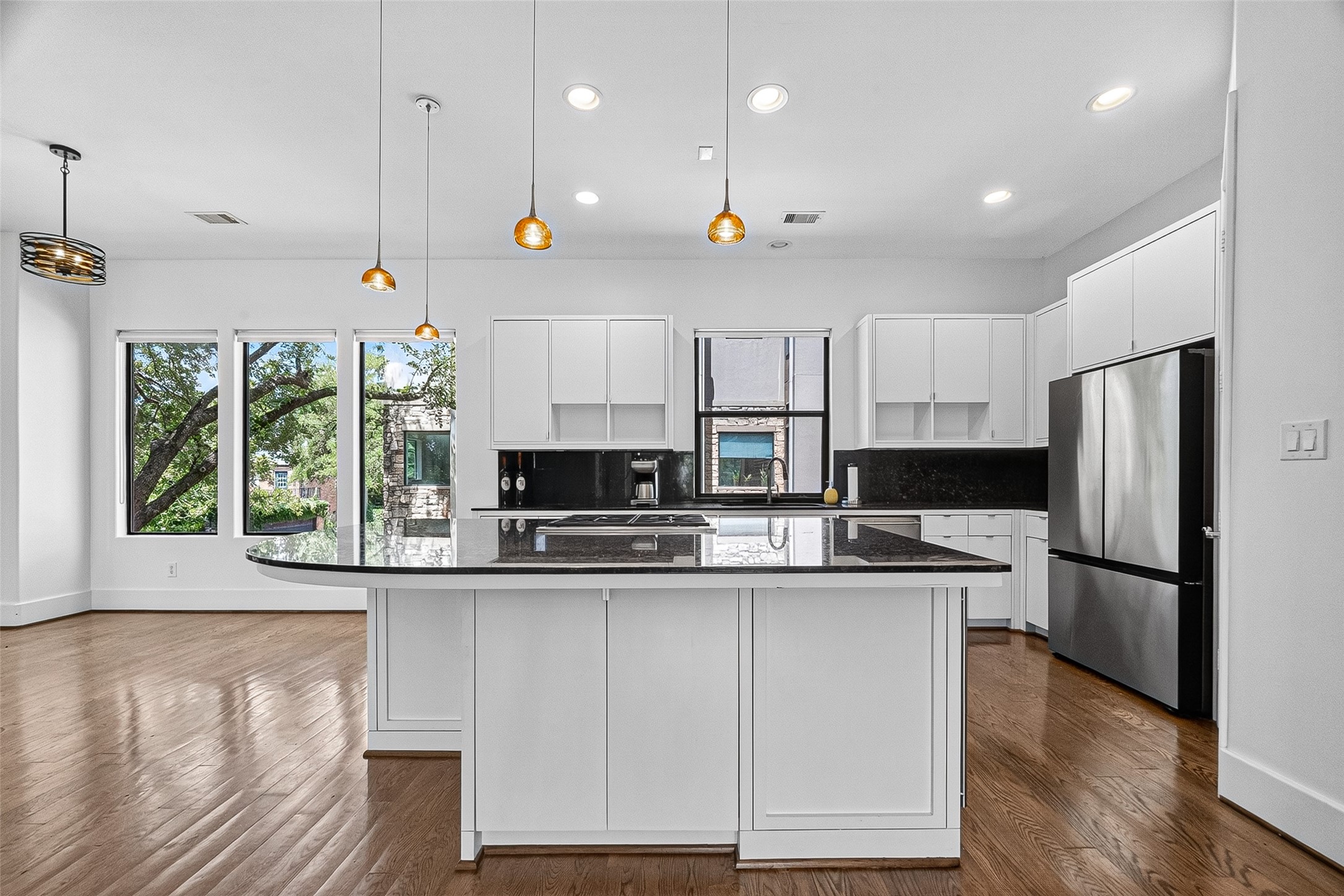 Bright and modern kitchen featuring sleek white cabinetry, a large center island with dark countertops, stainless steel appliances, and ample natural light from expansive windows.