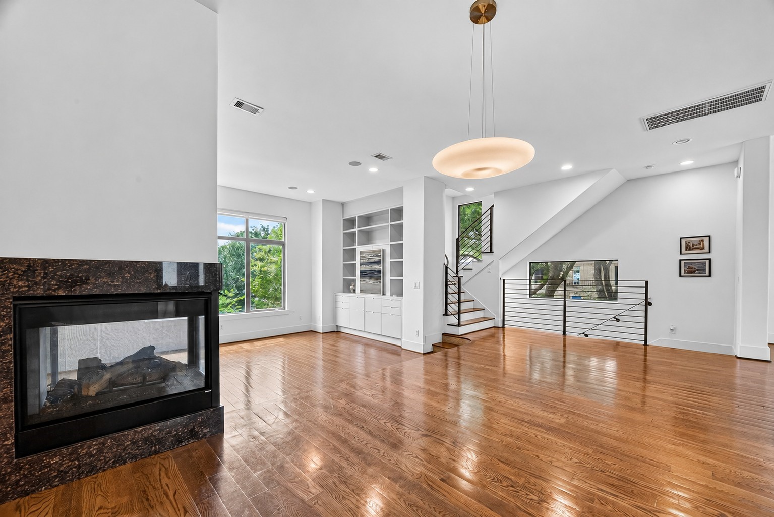 Elegant open-concept living space featuring rich hardwood floors, a modern gas fireplace, large windows for natural light, built-in shelving, and a sleek staircase with contemporary railing.