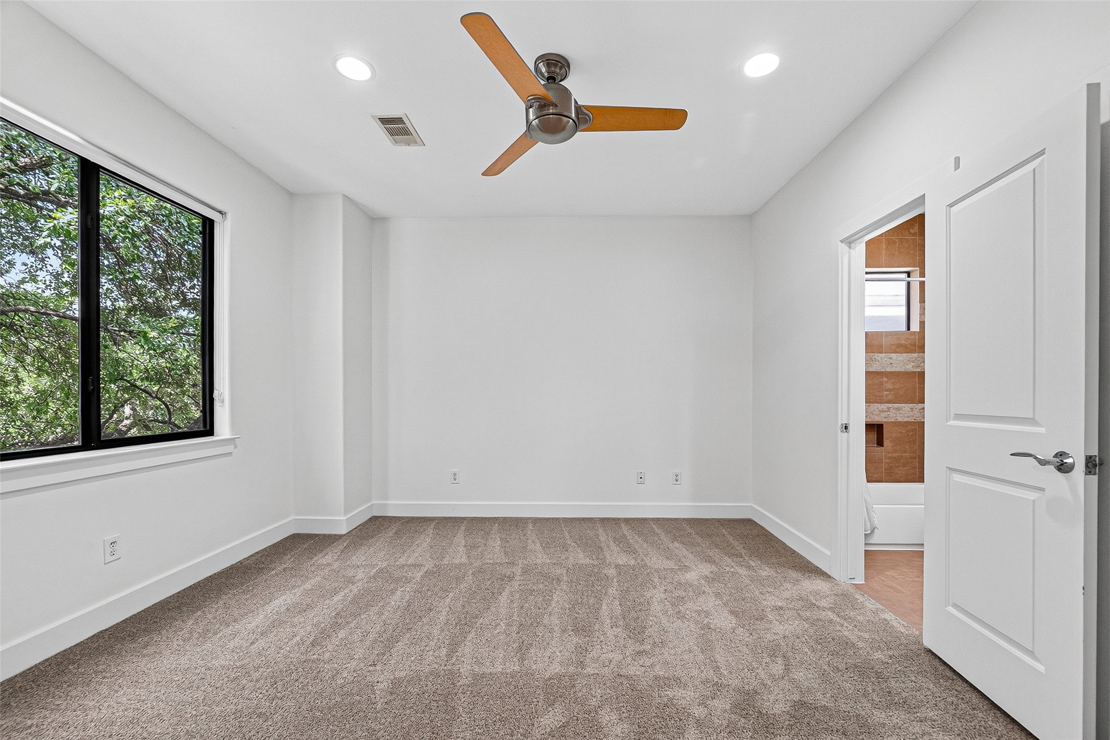 Spacious bedroom on second floor with plush carpeting, a modern ceiling fan, and large window providing natural light. Similar bedroom also on the first floor with en-suite bathroom.