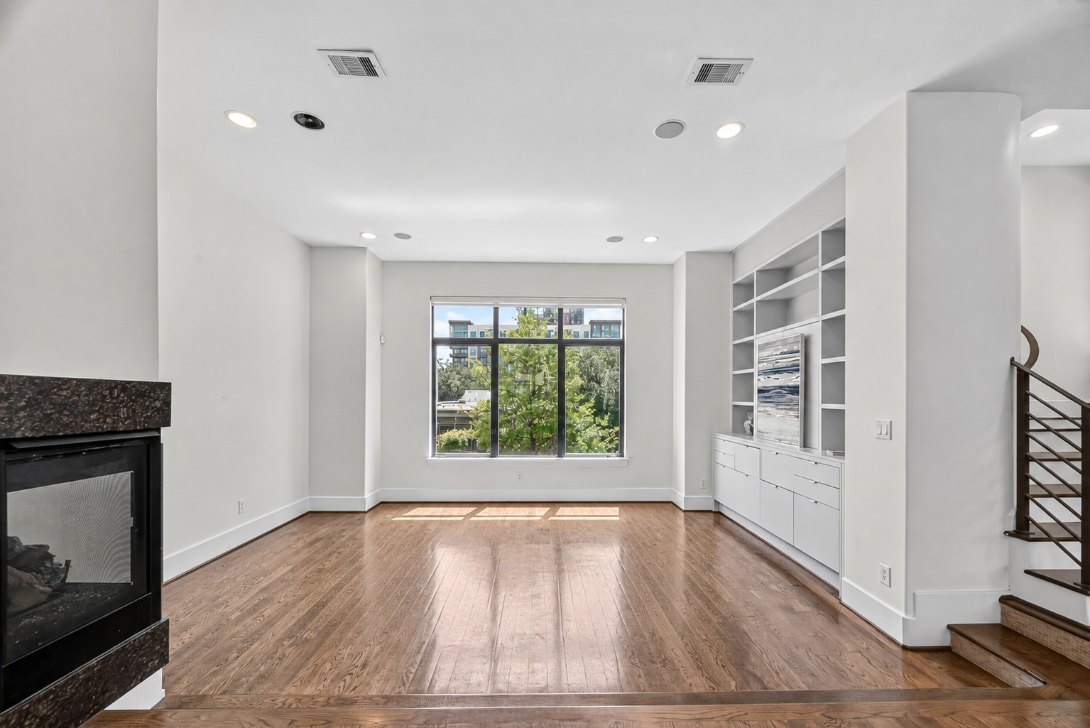 Bright and spacious living area with hardwood floors, a cozy fireplace, and built-in shelving, complemented by a large window offering natural light and views of greenery.