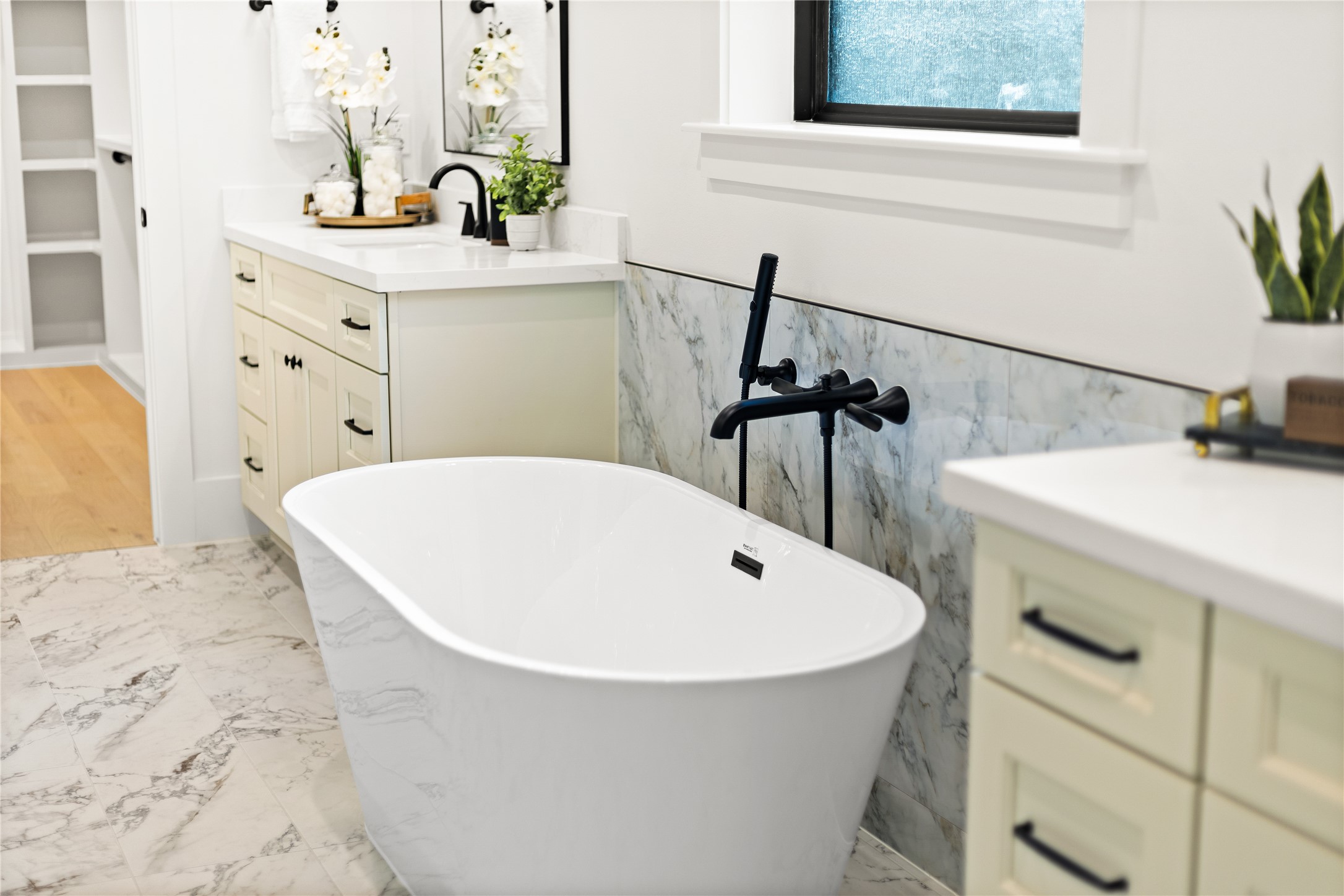 Unwind in your spa-inspired primary bath, where a sculptural freestanding soaking tub takes center stage against elegant marble tile. Striking matte black fixtures, including a floor-mounted tub filler and handheld sprayer, add a bold, modern contrast throughout the space. Dual vanities topped with quartz counters offer generous storage and prep space, while a frosted window brings in soft natural light and privacy.