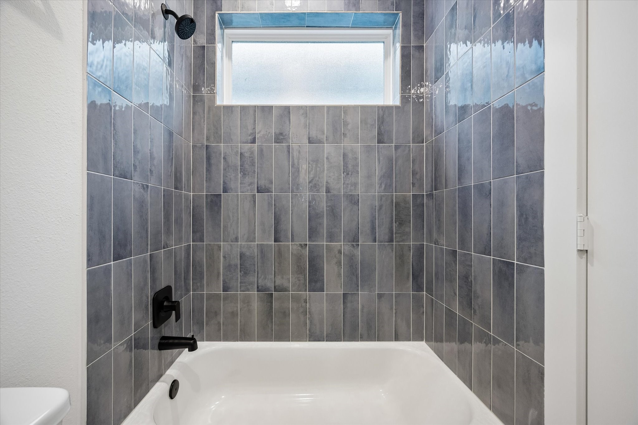 Modern bathroom with sleek gray tile walls, featuring a pristine bathtub and stylish black fixtures, illuminated by a frosted window for natural light.