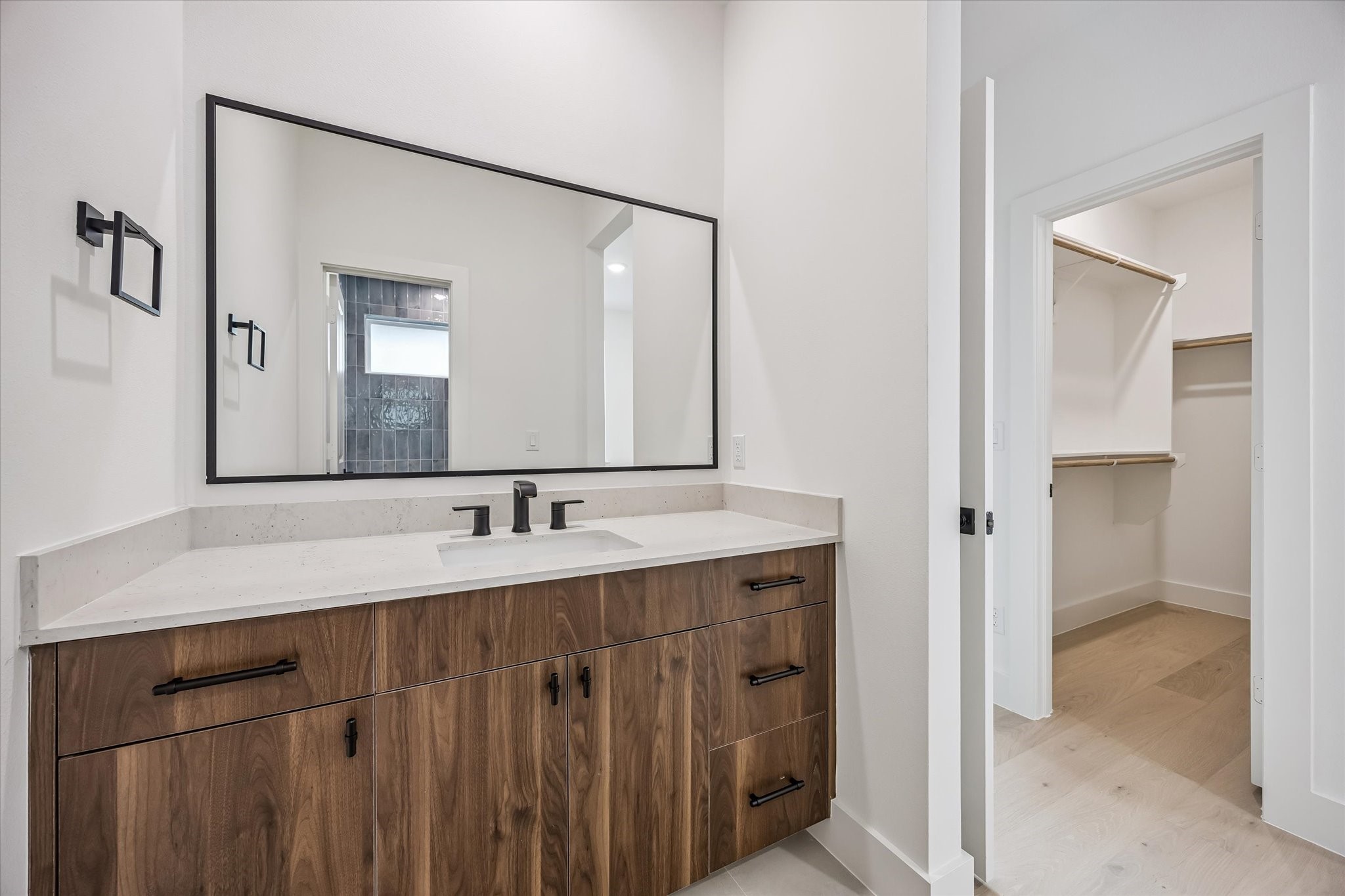 Modern bathroom featuring a sleek vanity with wood cabinetry, a large mirror, and black fixtures, leading to a spacious walk-in closet with ample shelving.