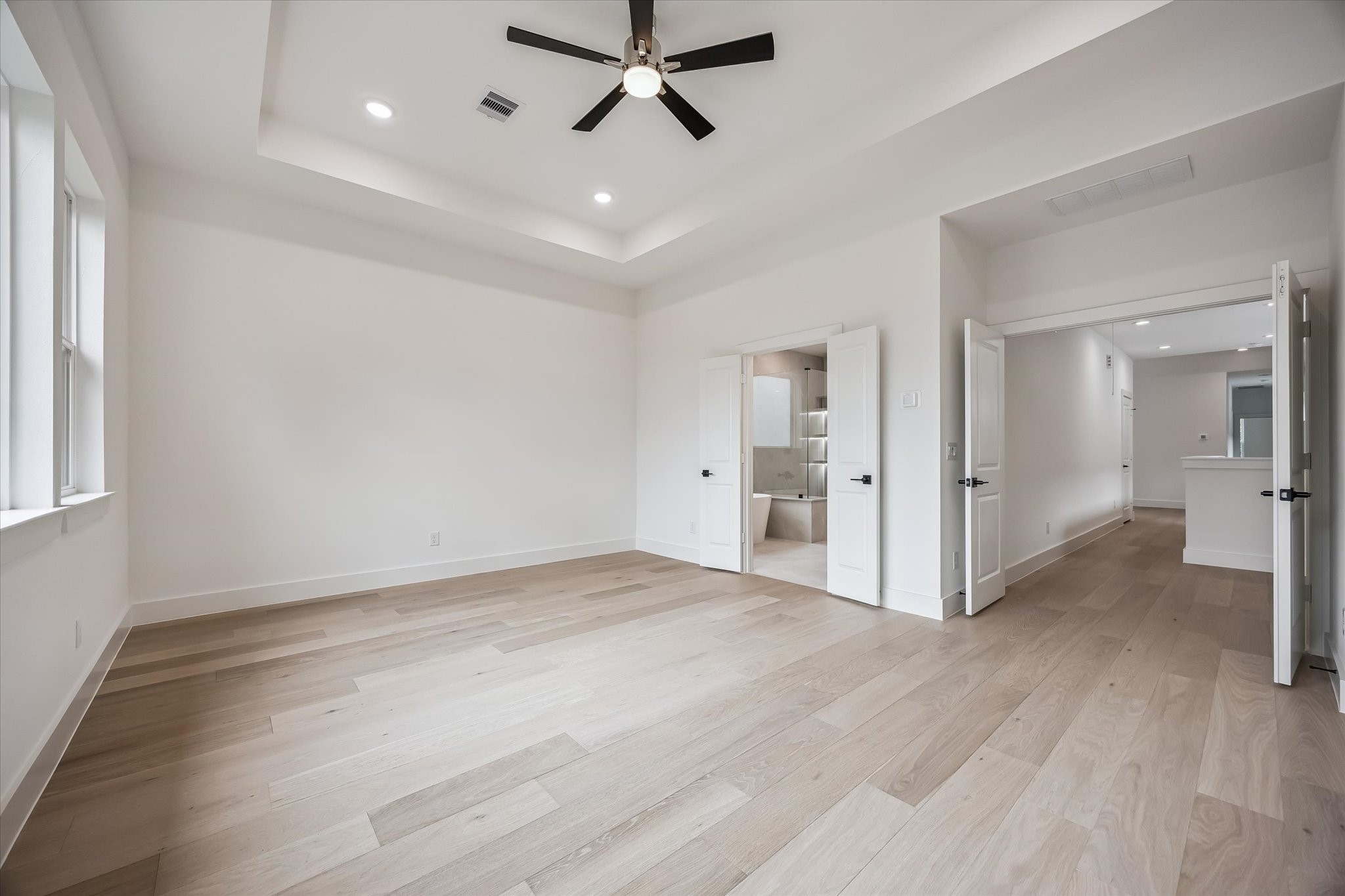 Spacious bedroom with elegant tray ceiling, recessed lighting, and light wood floors, leading to an en-suite bathroom through double doors.