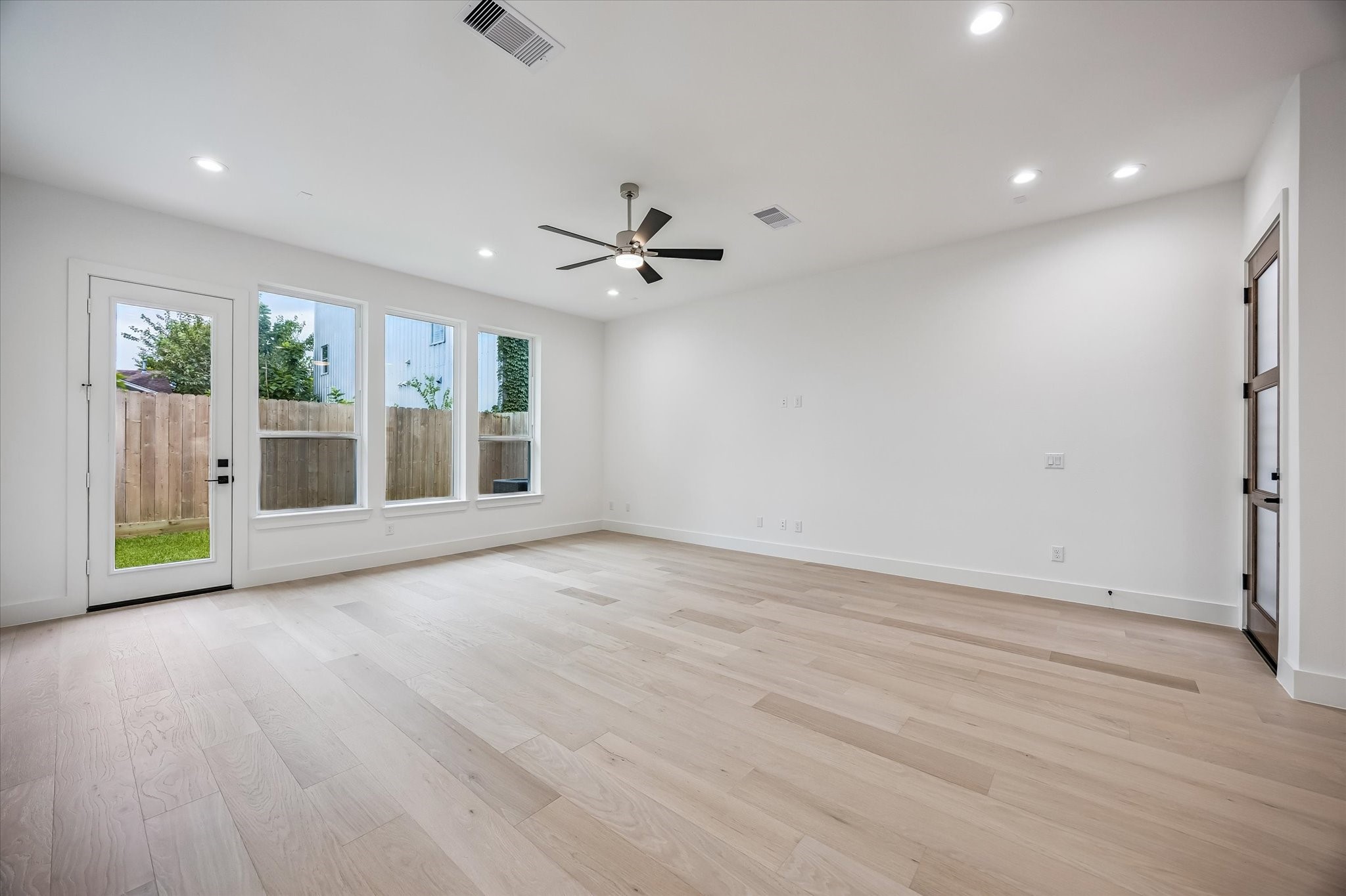 Spacious living area with light wood flooring, abundant natural light from large windows, and a modern ceiling fan, creating an inviting atmosphere. Spacious living area with light wood flooring, abundant natural light from large windows, and a modern ceiling fan, creating an inviting atmosphere.