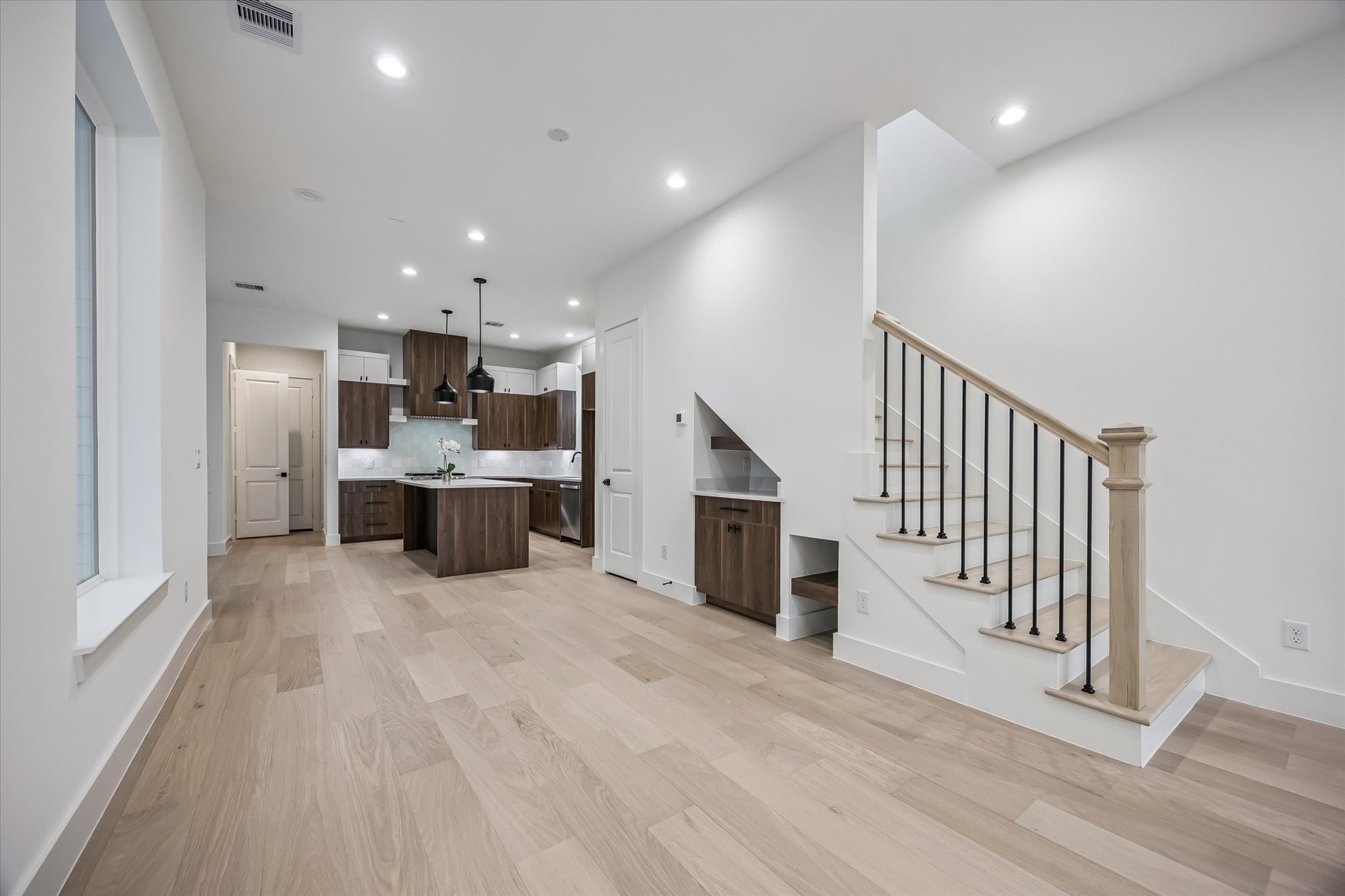 Modern open-concept living area featuring sleek wood flooring, a stylish kitchen with pendant lighting, and a contemporary staircase with black iron railings.