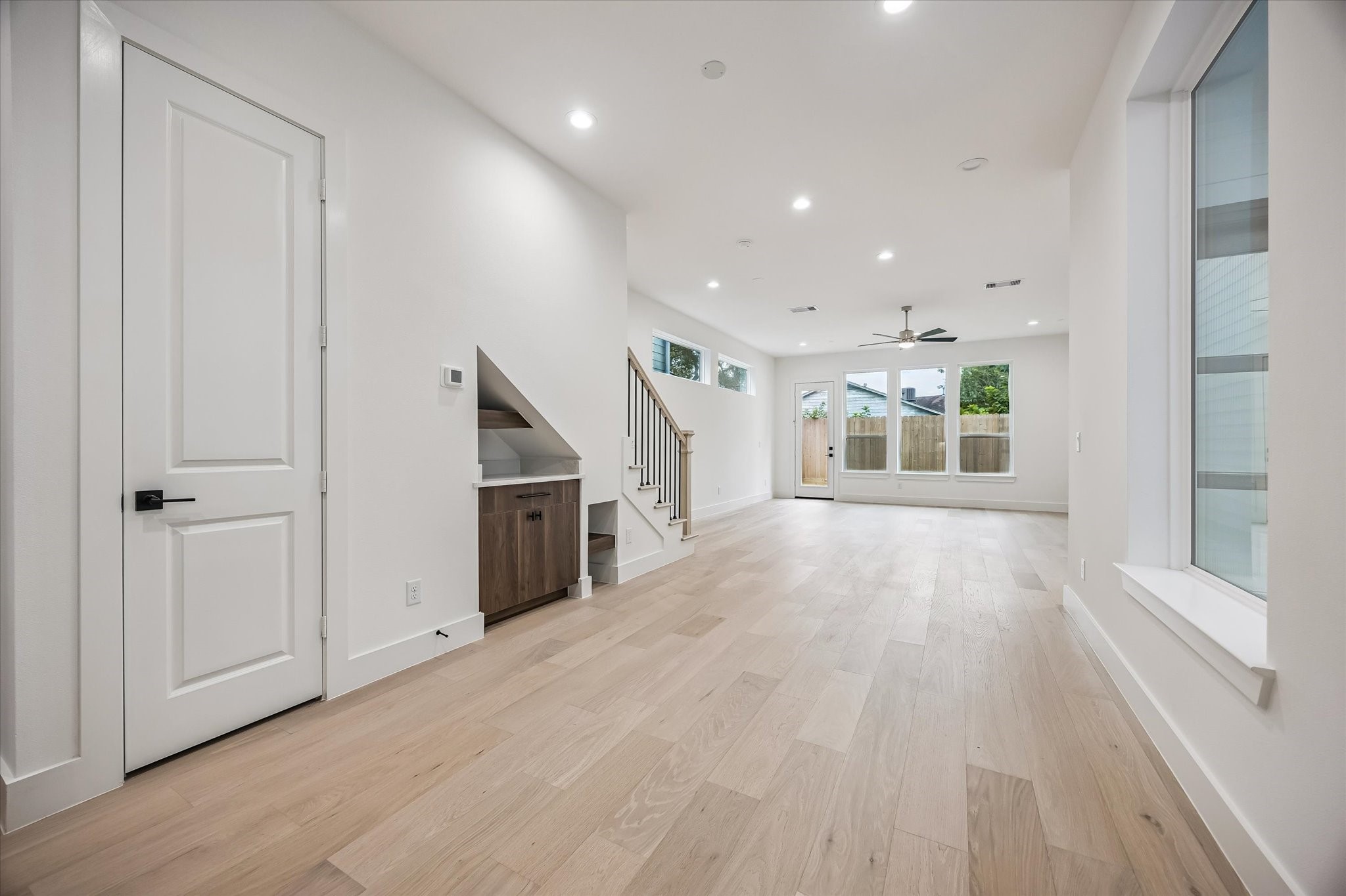 Spacious and bright living area with light wood floors, recessed lighting, and large windows leading to a backyard. Features a modern staircase and convenient built-in storage under the stairs.