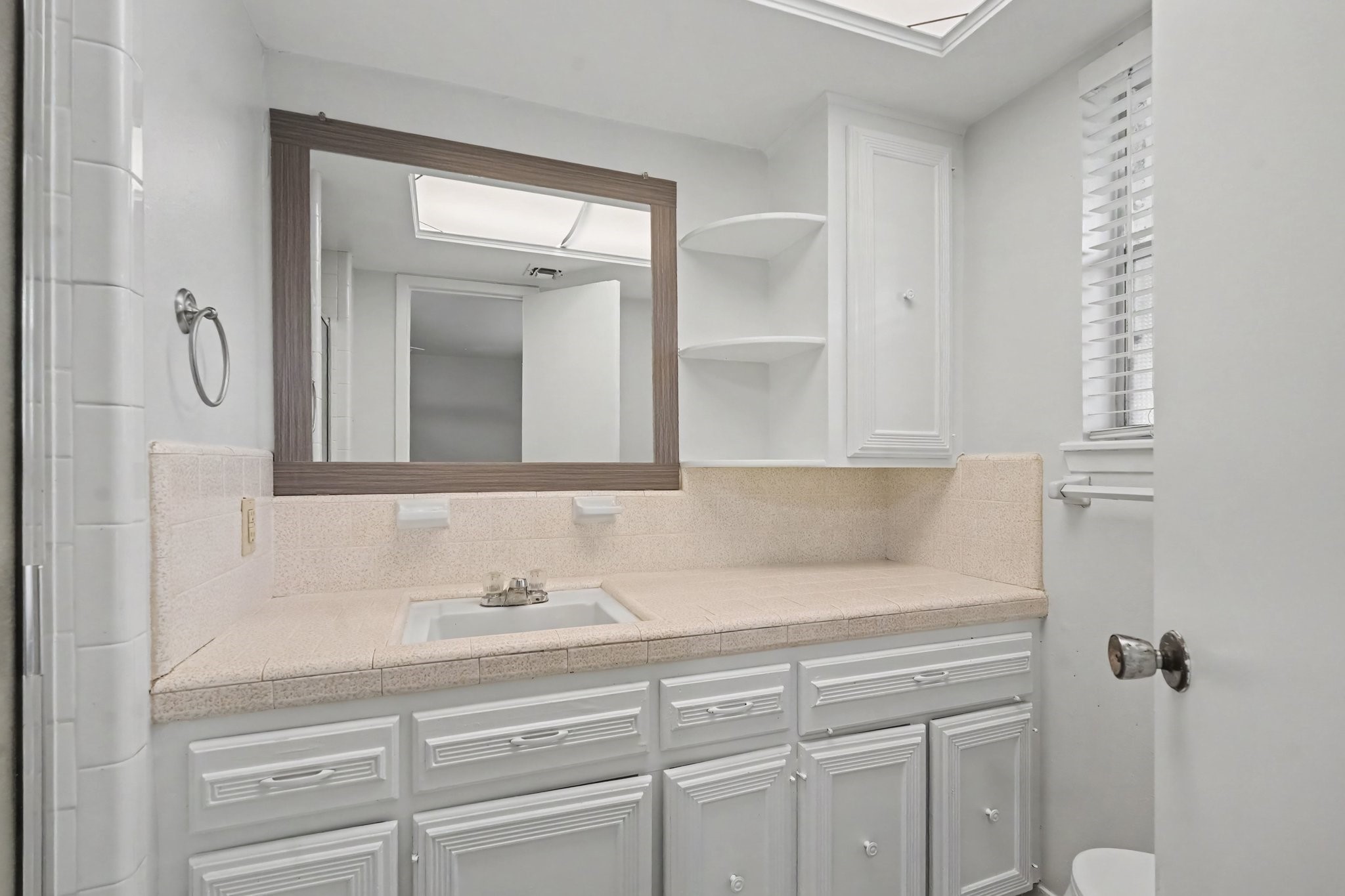 This ensuite bath is light and bright, featuring ample storage with built-in drawers and cabinets, convenient corner shelving, and a charming window that allows for plenty of natural light.