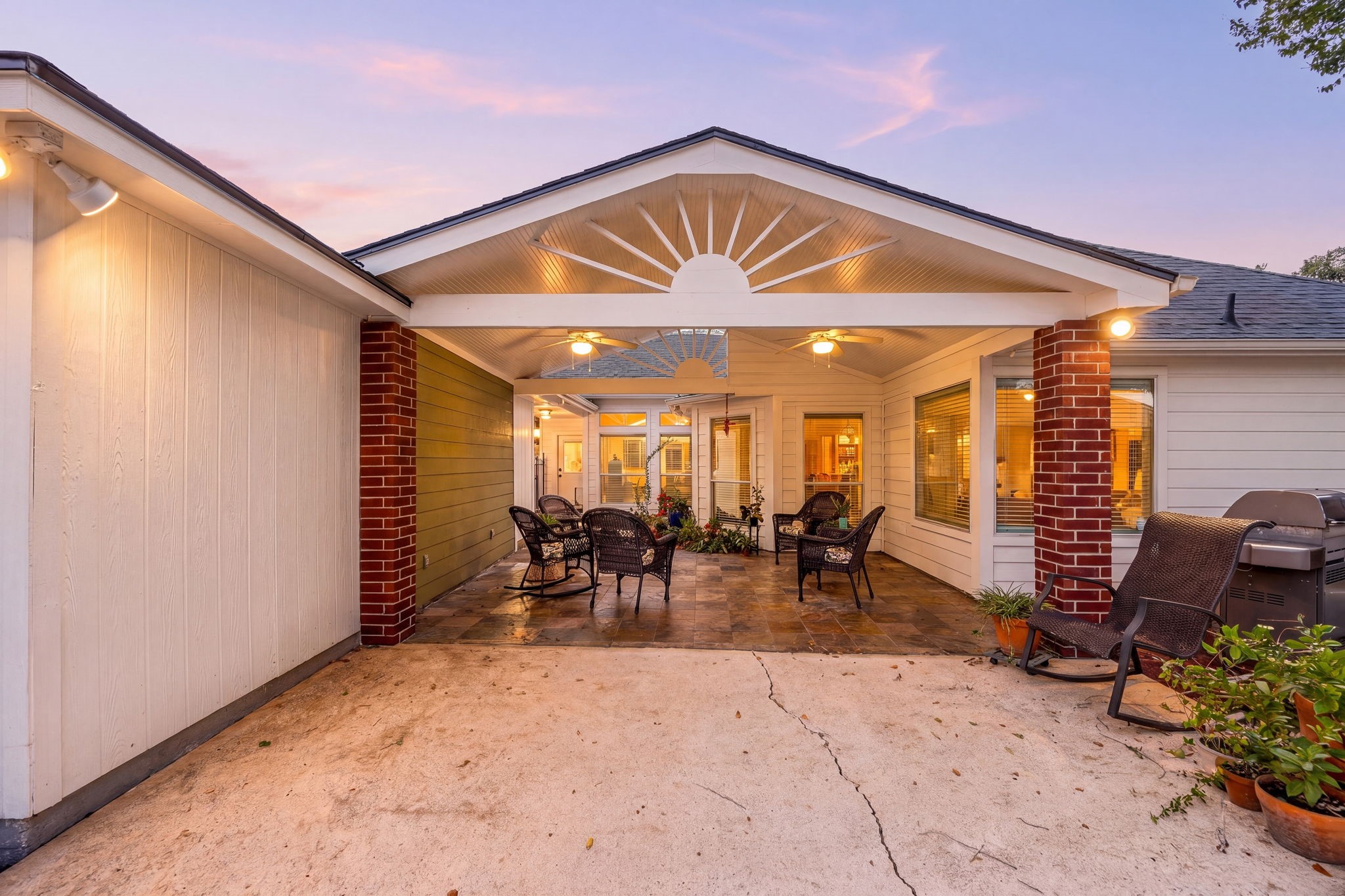 As evening falls, the covered patio glows beautifully, showcasing the tiled flooring, brick columns, and the additional concrete slab extending the outdoor living space.