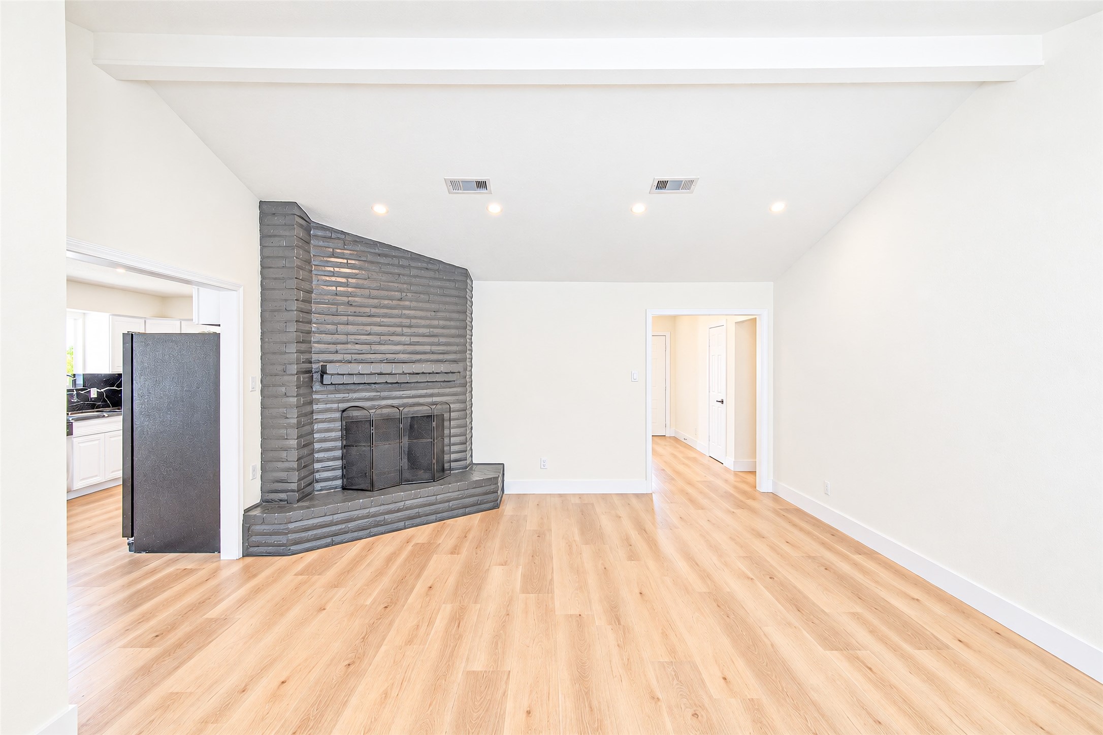 Spacious living area featuring a unique angled brick fireplace, high ceilings, and warm wood flooring, seamlessly connecting to the kitchen and hallway.