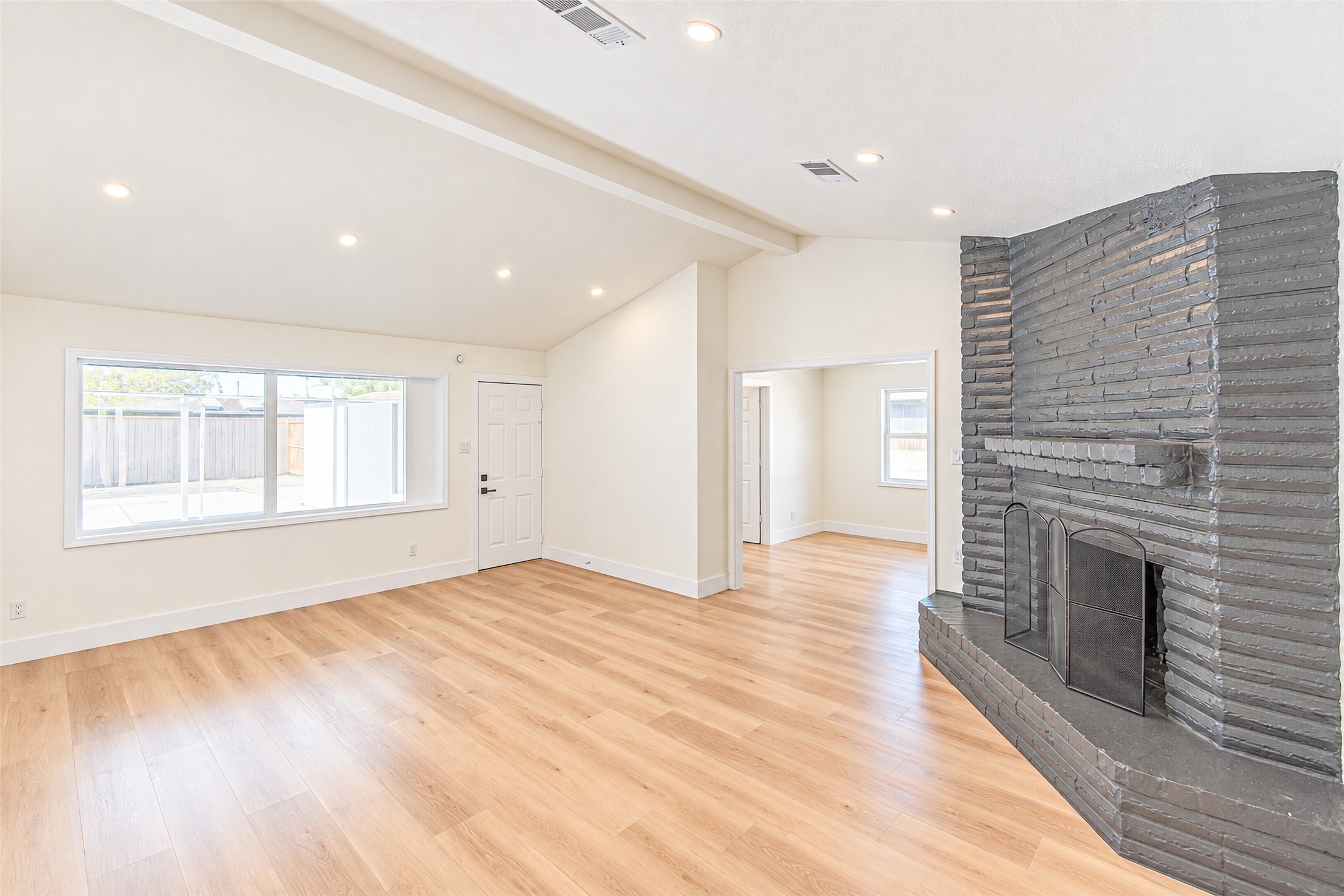 Bright and airy living room with vaulted ceilings, featuring a striking brick fireplace and sleek hardwood floors, complemented by large windows that flood the space with natural light.