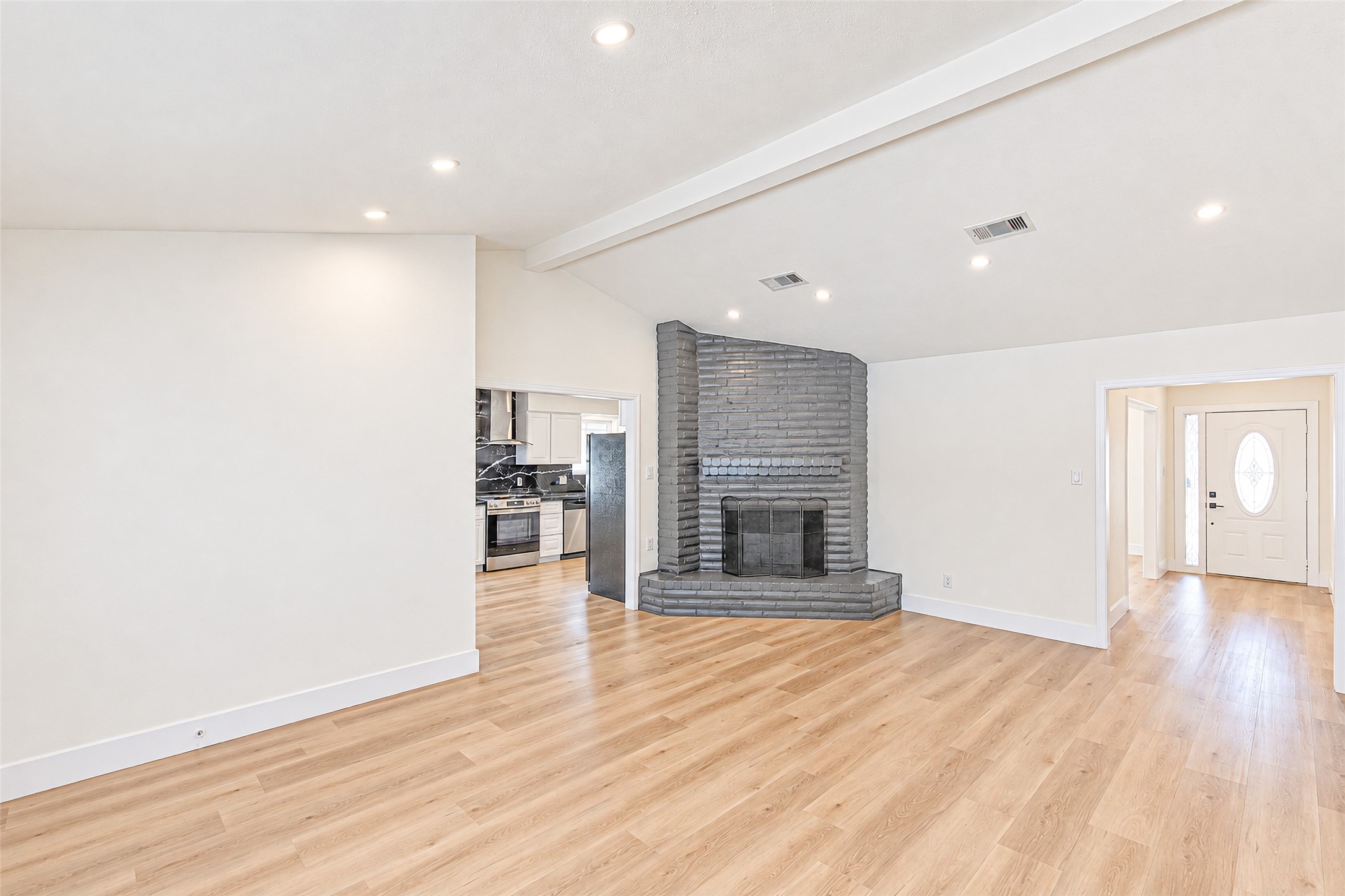 Spacious living area with sleek wood flooring, a striking gray brick fireplace, and recessed lighting, leading to a modern kitchen and bright entryway.