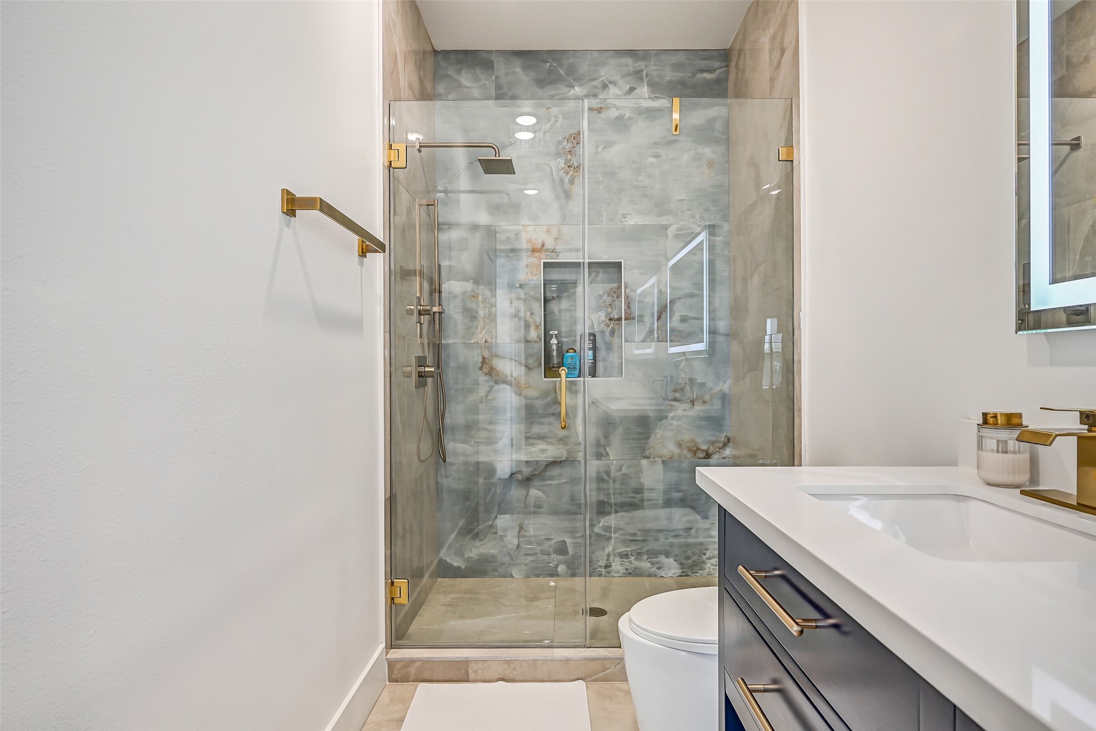 This bathroom features quartz countertops, double sinks, gold fixtures, and a walk-in shower with a waterfall showerhead and stunning large tile surround.