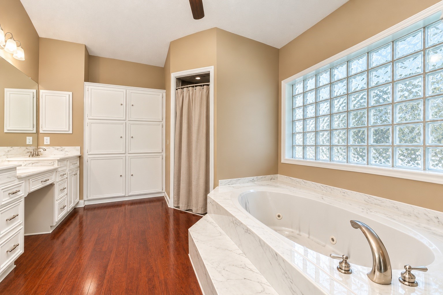 Luxurious master bathroom featuring a spacious corner jacuzzi tub, elegant glass block window, and dual-sink vanity with ample storage.
