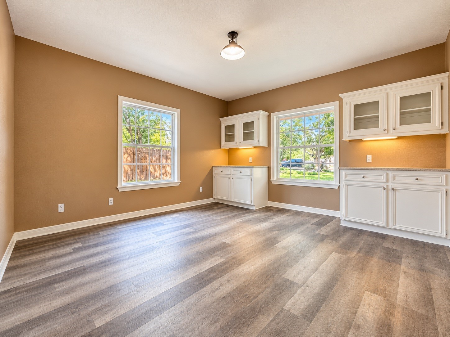 Bright and inviting dining room with warm-toned walls, featuring large windows for natural light, elegant white cabinetry with glass fronts, and stylish wood-look flooring.