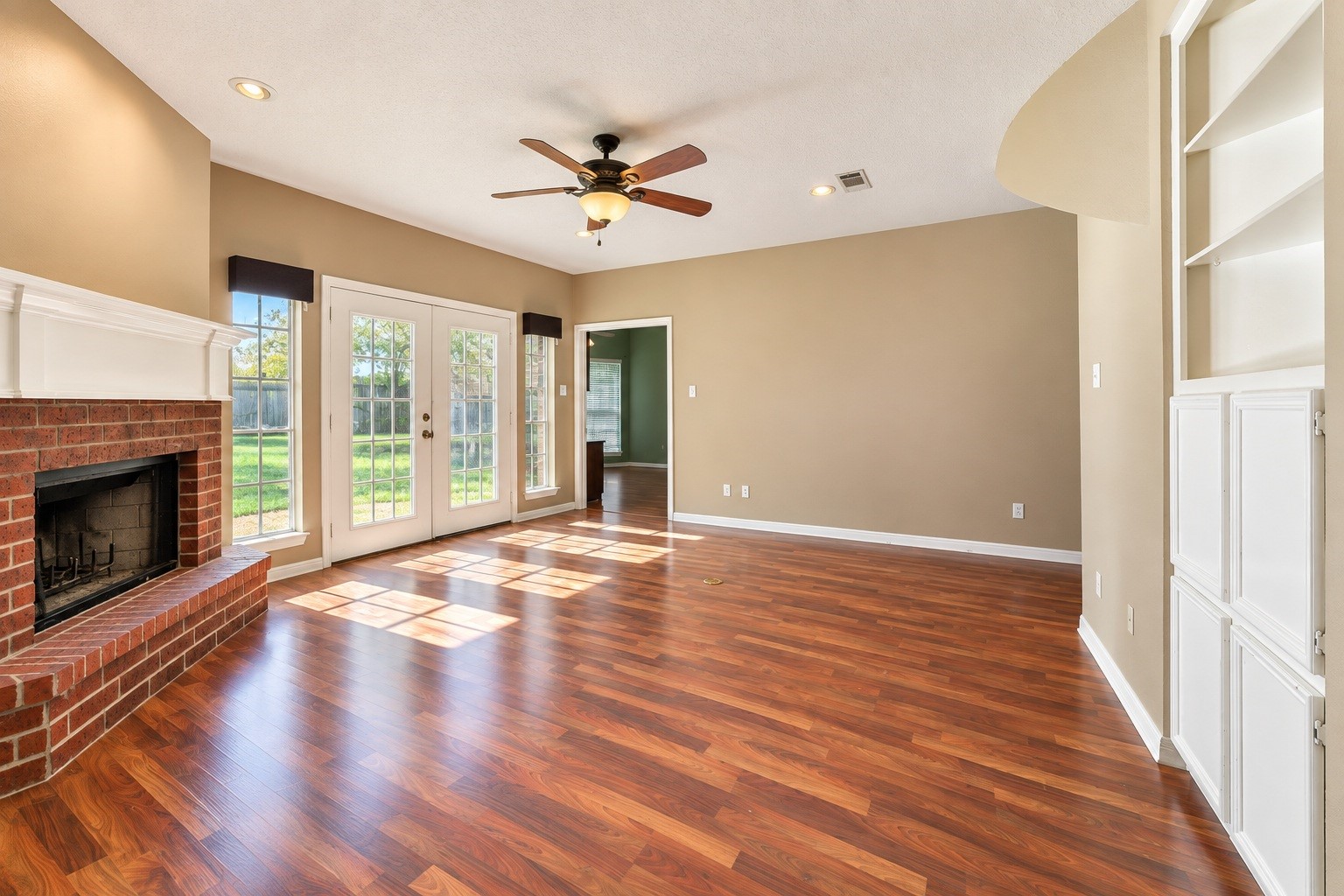 Inviting living room with rich wood-look floors, a cozy brick fireplace, and abundant natural light streaming through French doors that open to a lush backyard.