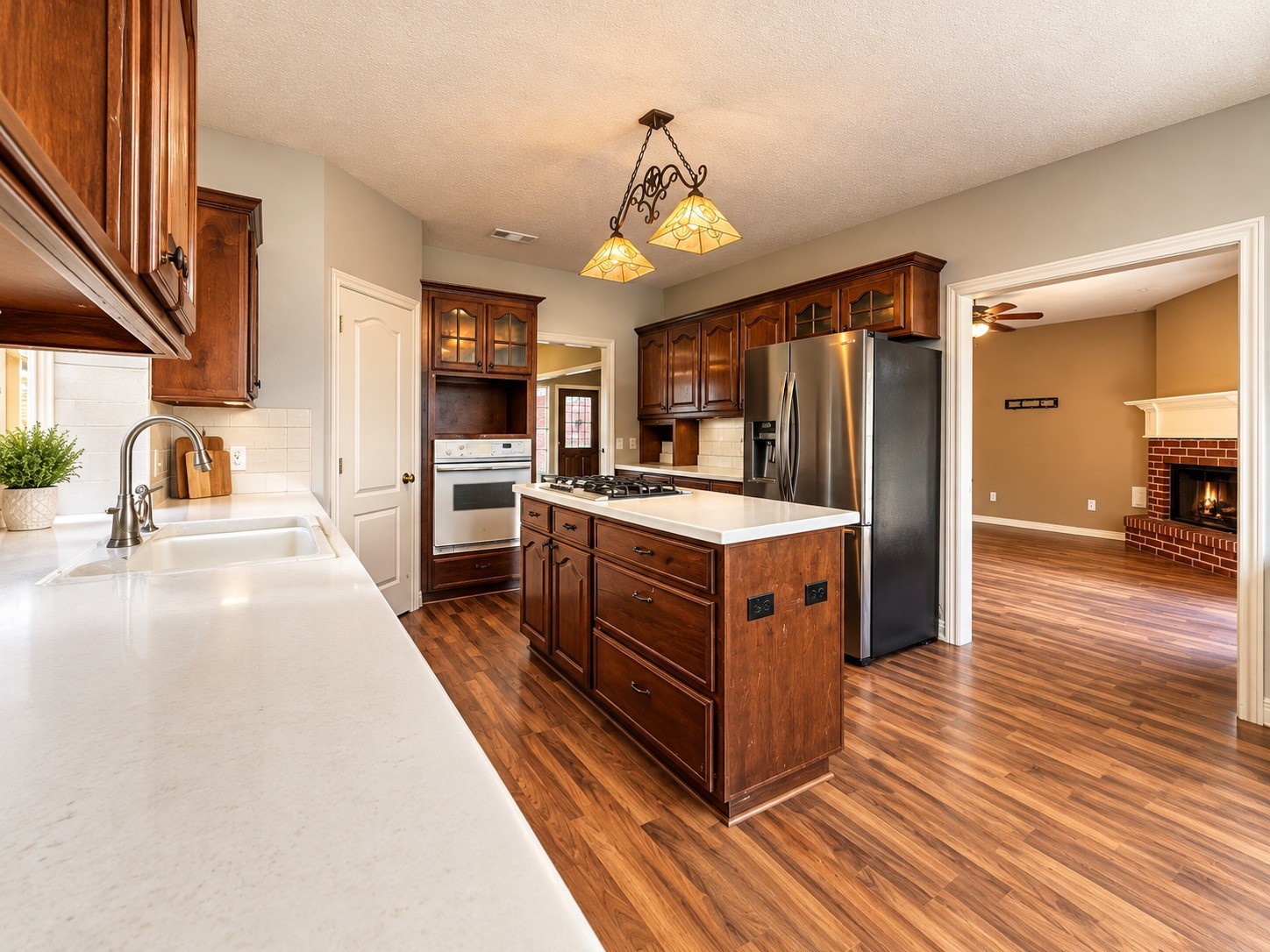 Spacious kitchen with rich wood cabinetry, modern stainless steel refrigerator, and a central island, seamlessly flowing into a cozy living room with a warm fireplace.
