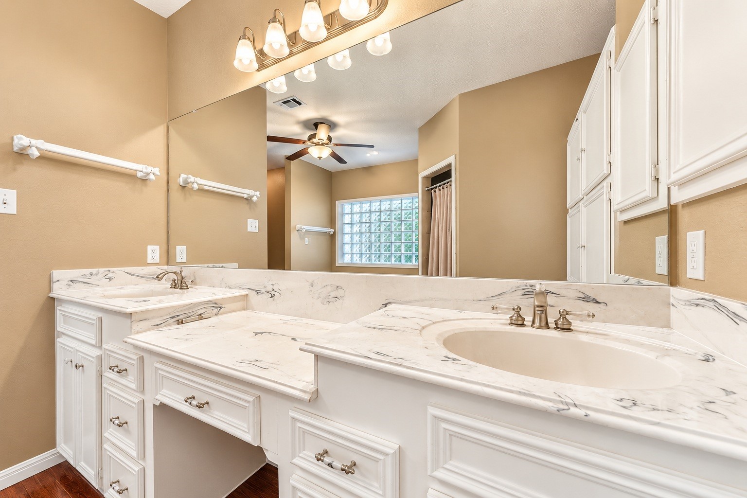 Elegant master bathroom featuring dual vanities with marble countertops, ample cabinet storage, and a large mirror, complemented by warm lighting and a ceiling fan.