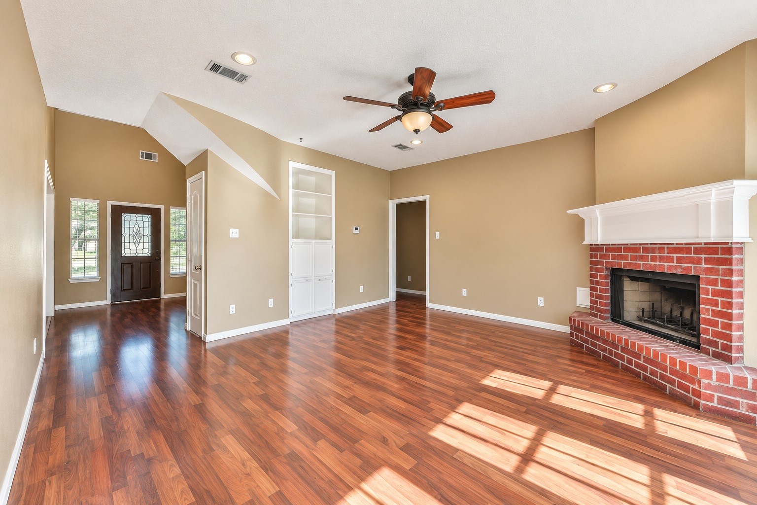 Spacious living room with rich wood-look floors, a cozy brick fireplace, and built-in shelving. Natural light pours in through large windows, complementing the ceiling fan and elegant entryway.
